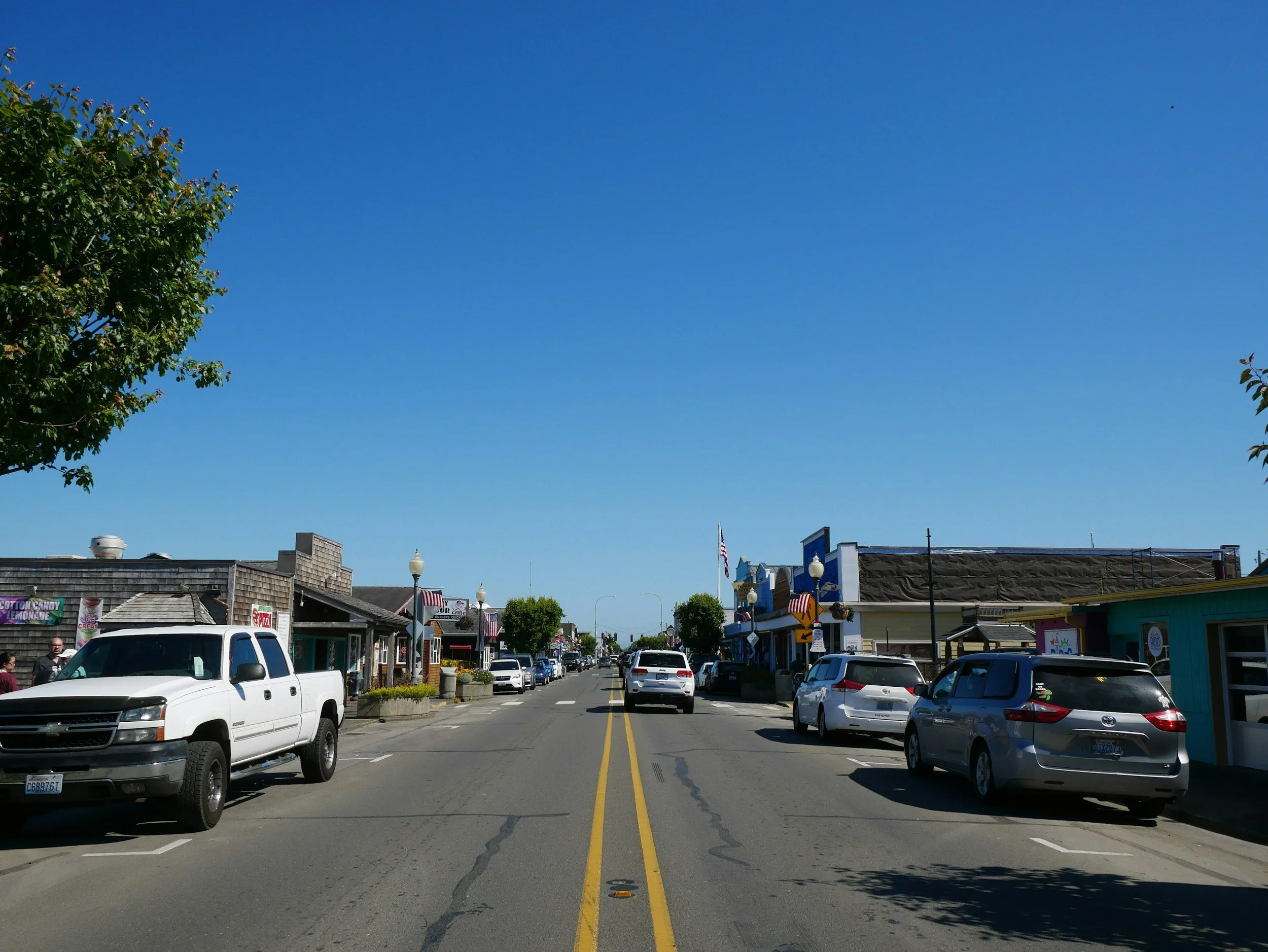 A small town street scene with parked cars on both sides, buildings with shops, and a clear blue sky with green trees visible on the left.