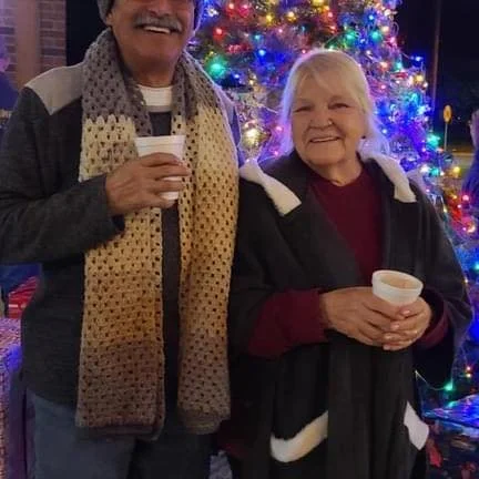 Victor Lemos and Kay Lemos, a man and an elderly woman stand in front of a decorated Christmas tree, both holding cups and smiling.