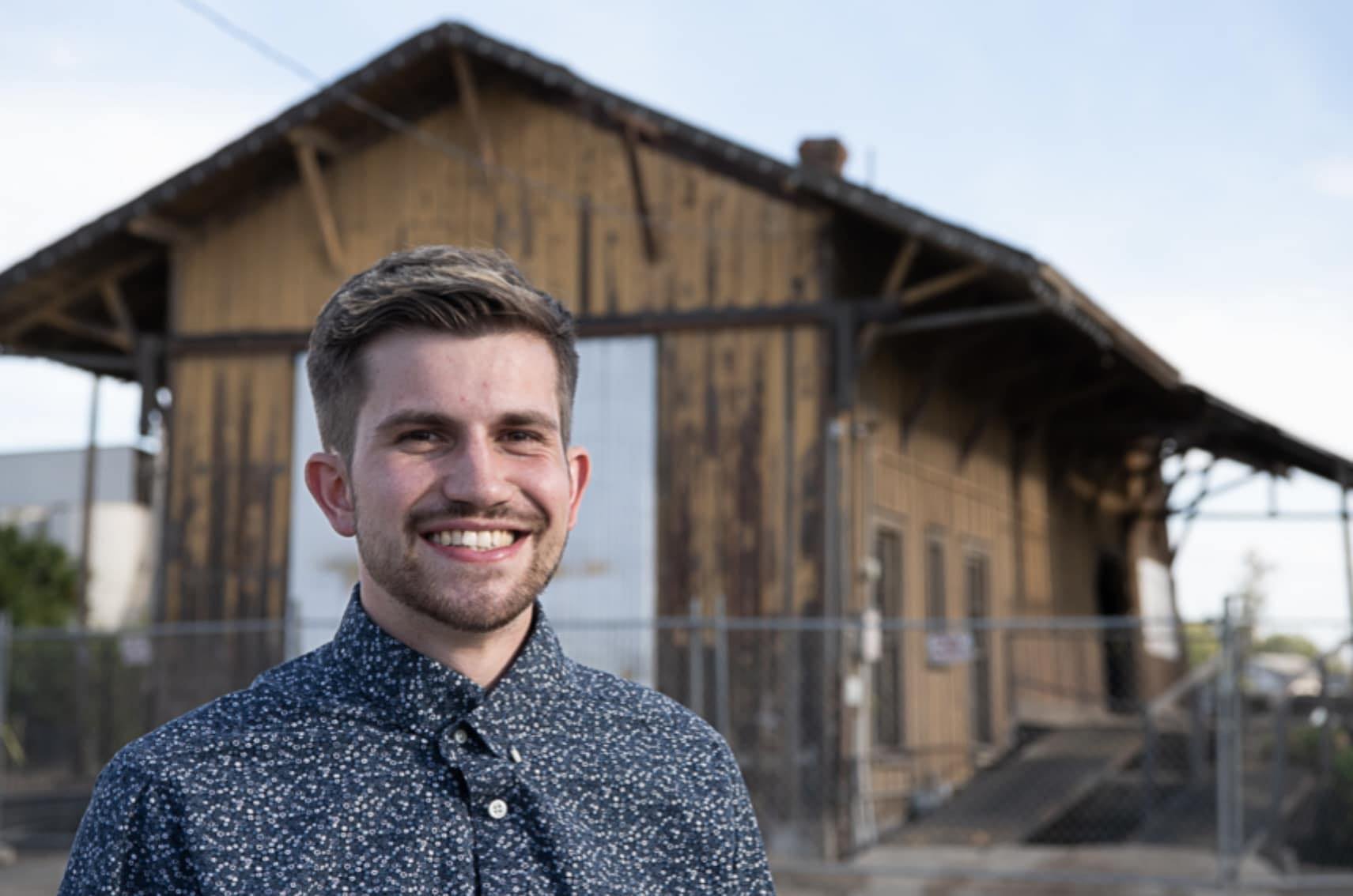 Portrait of Elijah Rodriguez, a smiling young man with short brown hair, wearing a dark patterned shirt, standing outdoors in front of the Arbuckle Train Depot, a rustic wooden building with clear skies in the background.