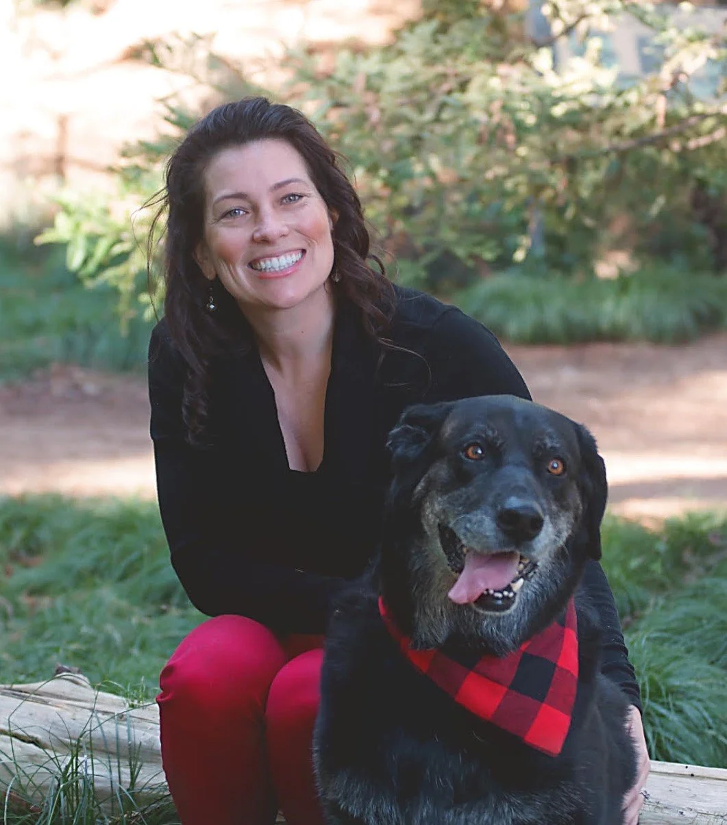 Lacey Gimple, a woman with dark hair smiling outdoors, sitting on the grass with her black and gray dog Cooper wearing a red bandana.