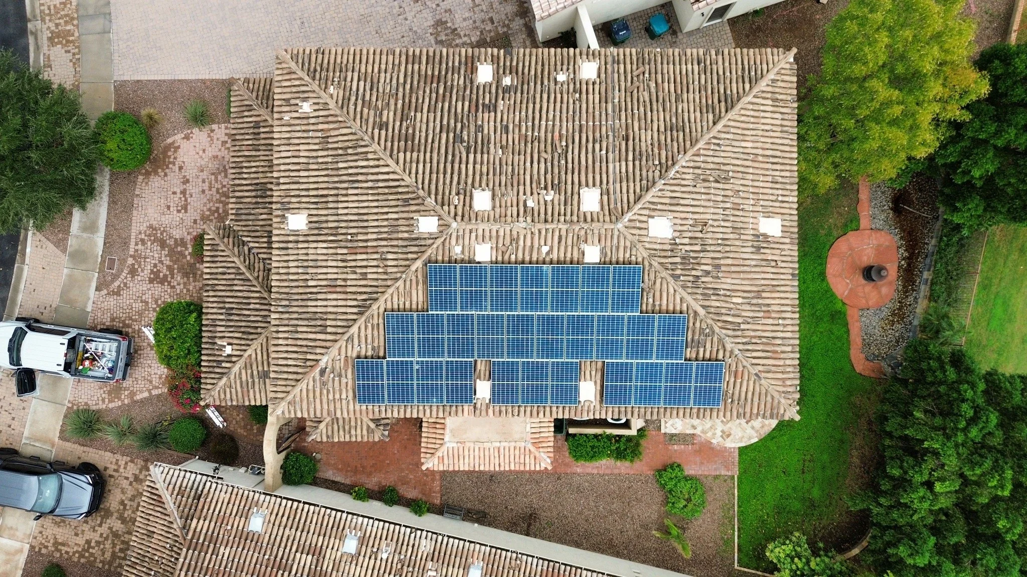 An aerial view of a house with a tiled roof installed with solar panels, surrounded by trees, greenery, a driveway, and parked vehicles.