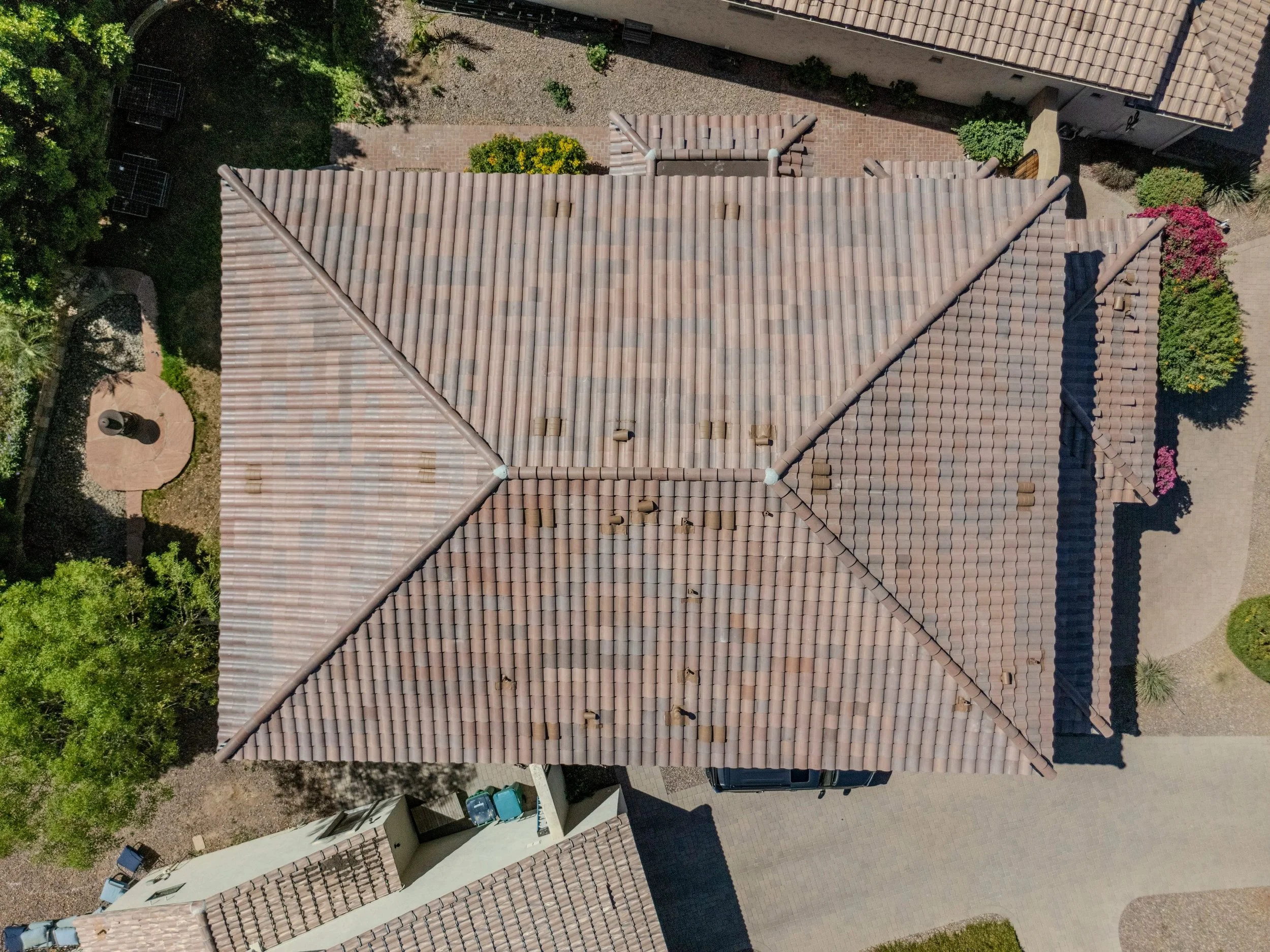 Aerial view of residential rooftops with beige tile roofs, surrounding trees, and a small backyard with a paved area, bushes, and a garden.