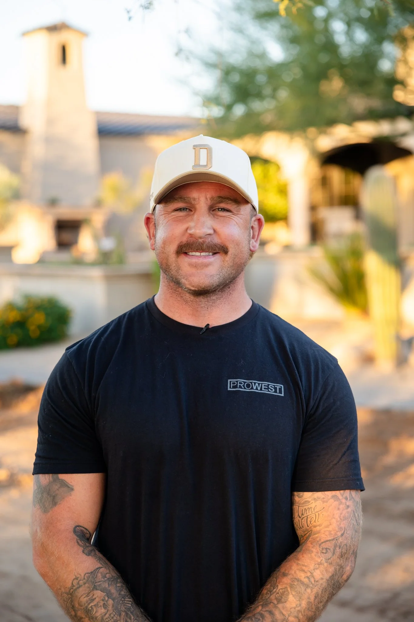 A man smiling outdoors wearing a white baseball cap and a black T-shirt with a logo, standing in front of a blurred background with trees and buildings, in warm lighting.
