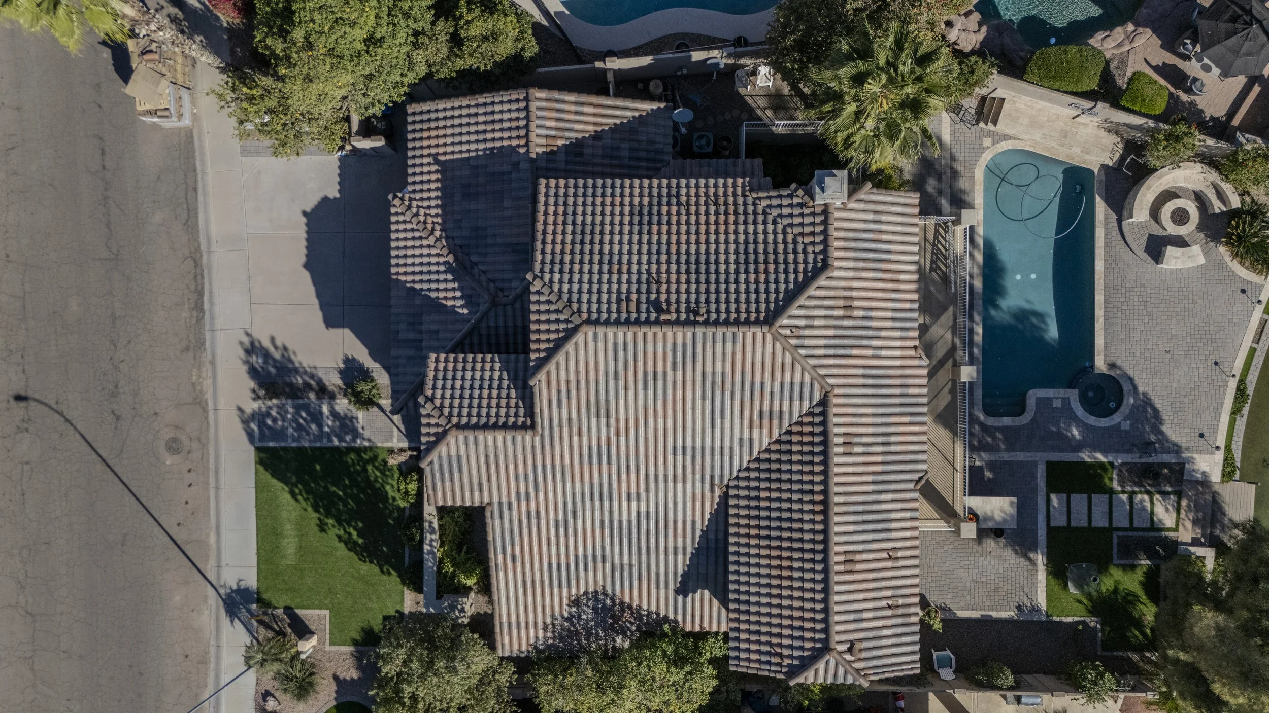 An aerial view of a house with a tiled roof, a pool, and surrounding landscaping, including trees and bushes.