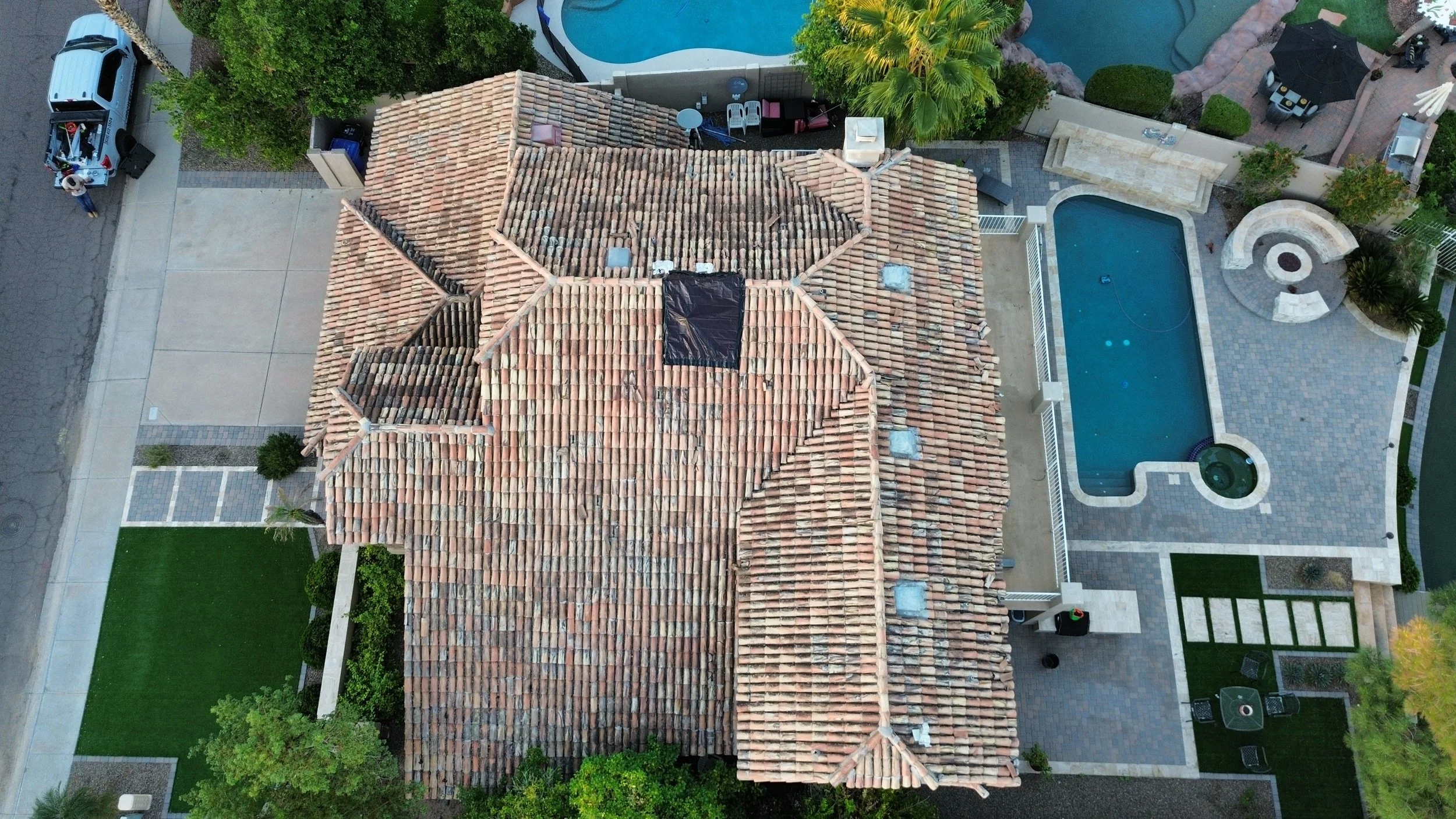 An aerial view of a house with a tiled roof surrounded by green trees, a swimming pool with a water feature, outdoor seating, and a backyard patio.