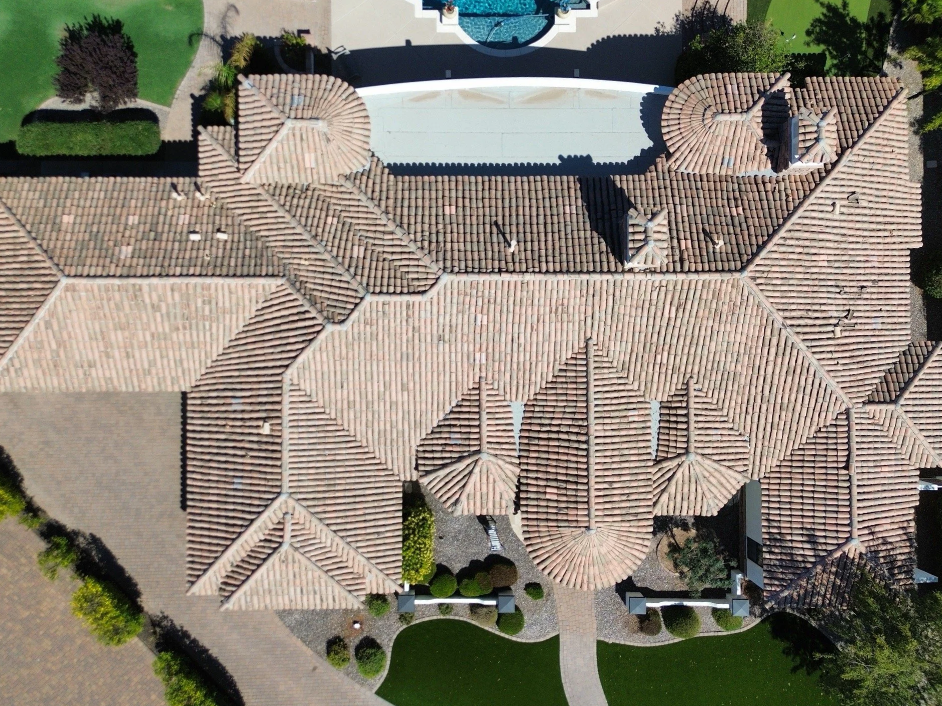An aerial view of a large house with a red tile roof, surrounded by landscaped gardens and walking paths. The house has multiple turrets and chimneys, and there is a swimming pool in the backyard.