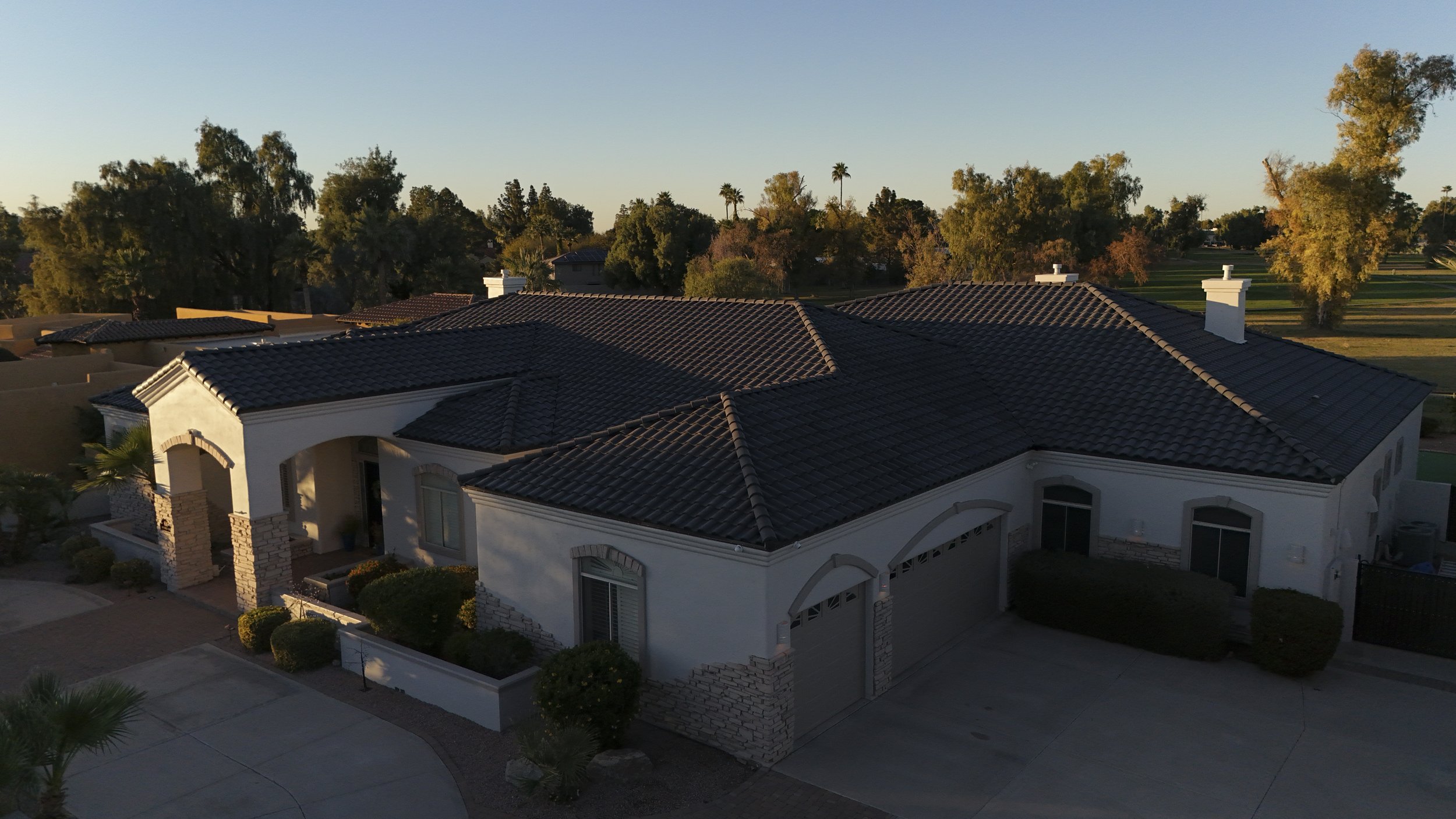 Aerial view of a white house with a dark tiled roof, surrounded by trees and a small front yard, during sunset.