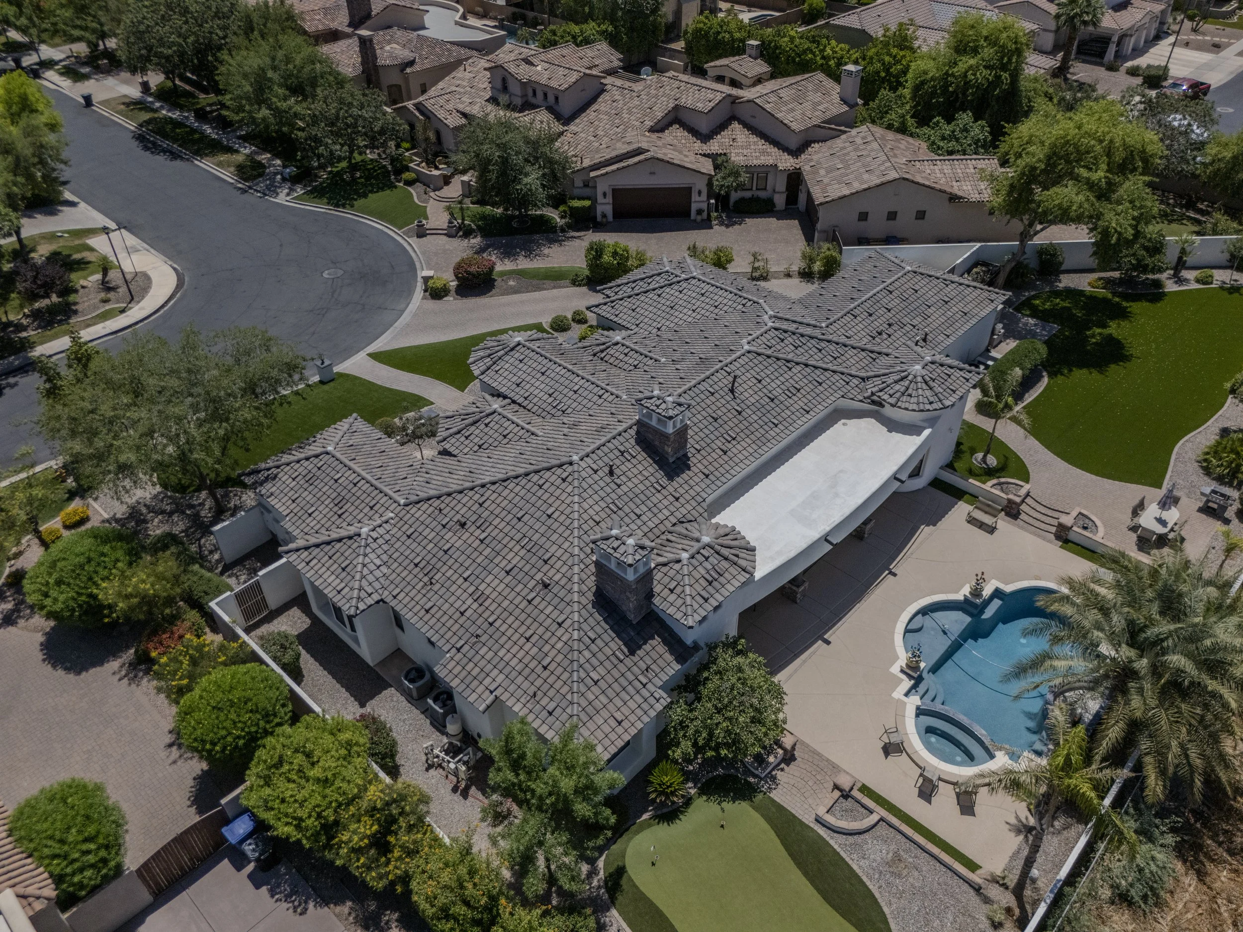 Aerial view of a residential neighborhood with large houses, tiled roofs, green lawns, trees, a swimming pool, paved walkways, and a curved street.