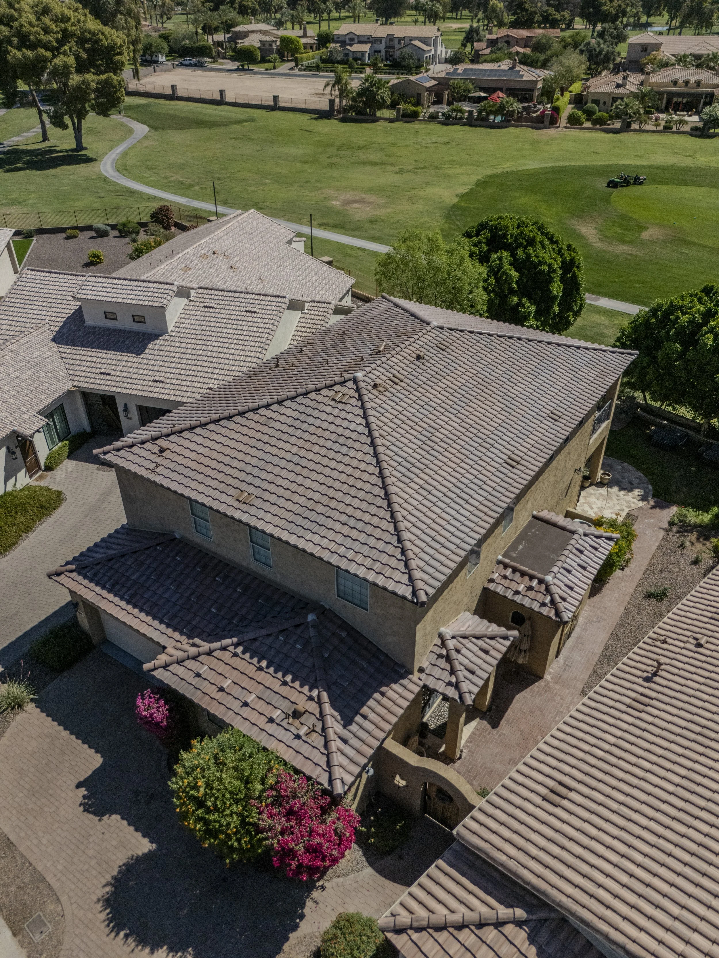 An aerial view of a residential neighborhood featuring two houses with tiled roofs, a wrapping driveway with landscaped shrubbery, trees, and flowering plants, with a large grassy park and other houses in the background.