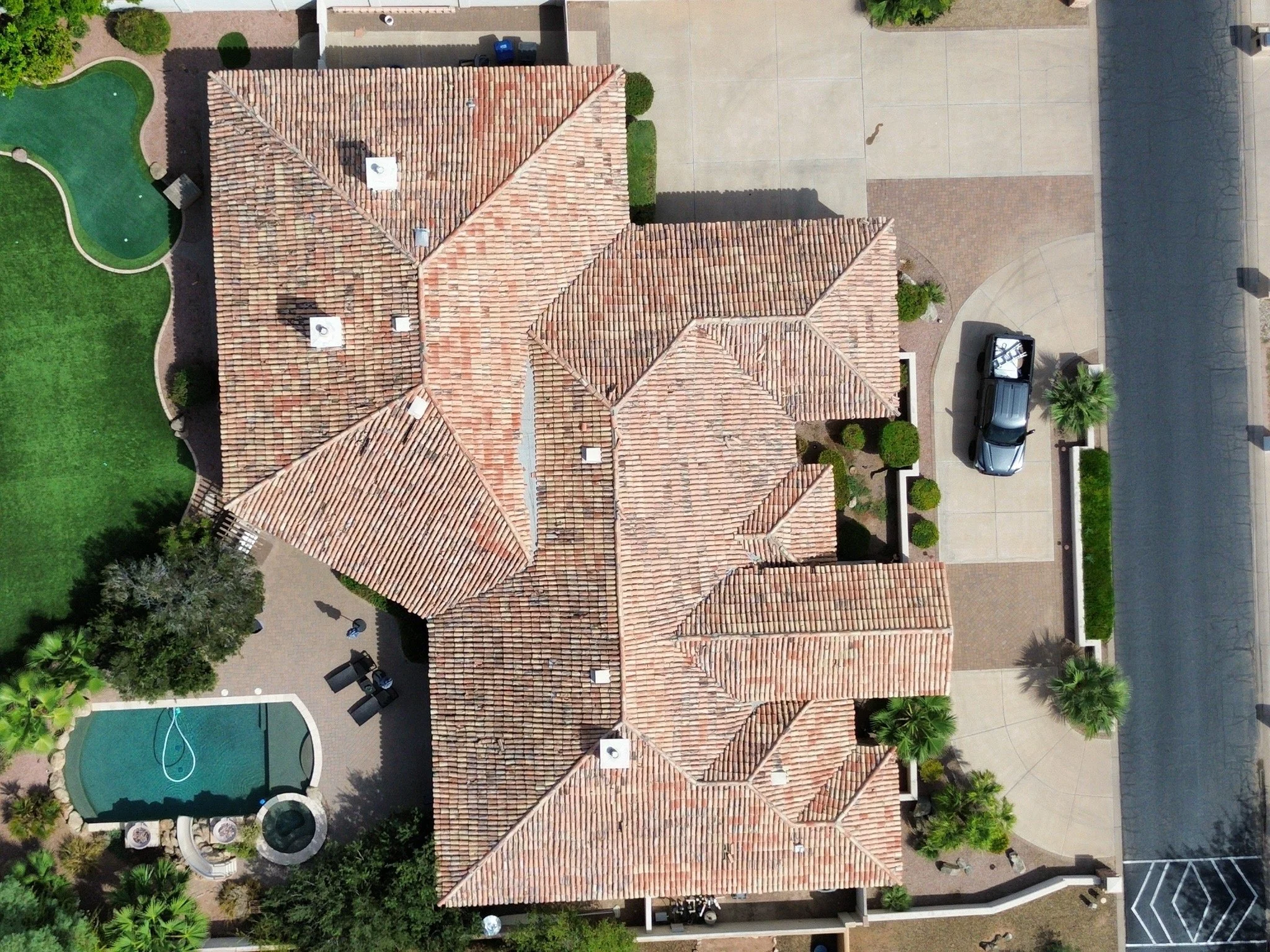Aerial view of a house with a terracotta tile roof, surrounded by manicured greenery, a swimming pool, and a concrete driveway with a black car parked on it.