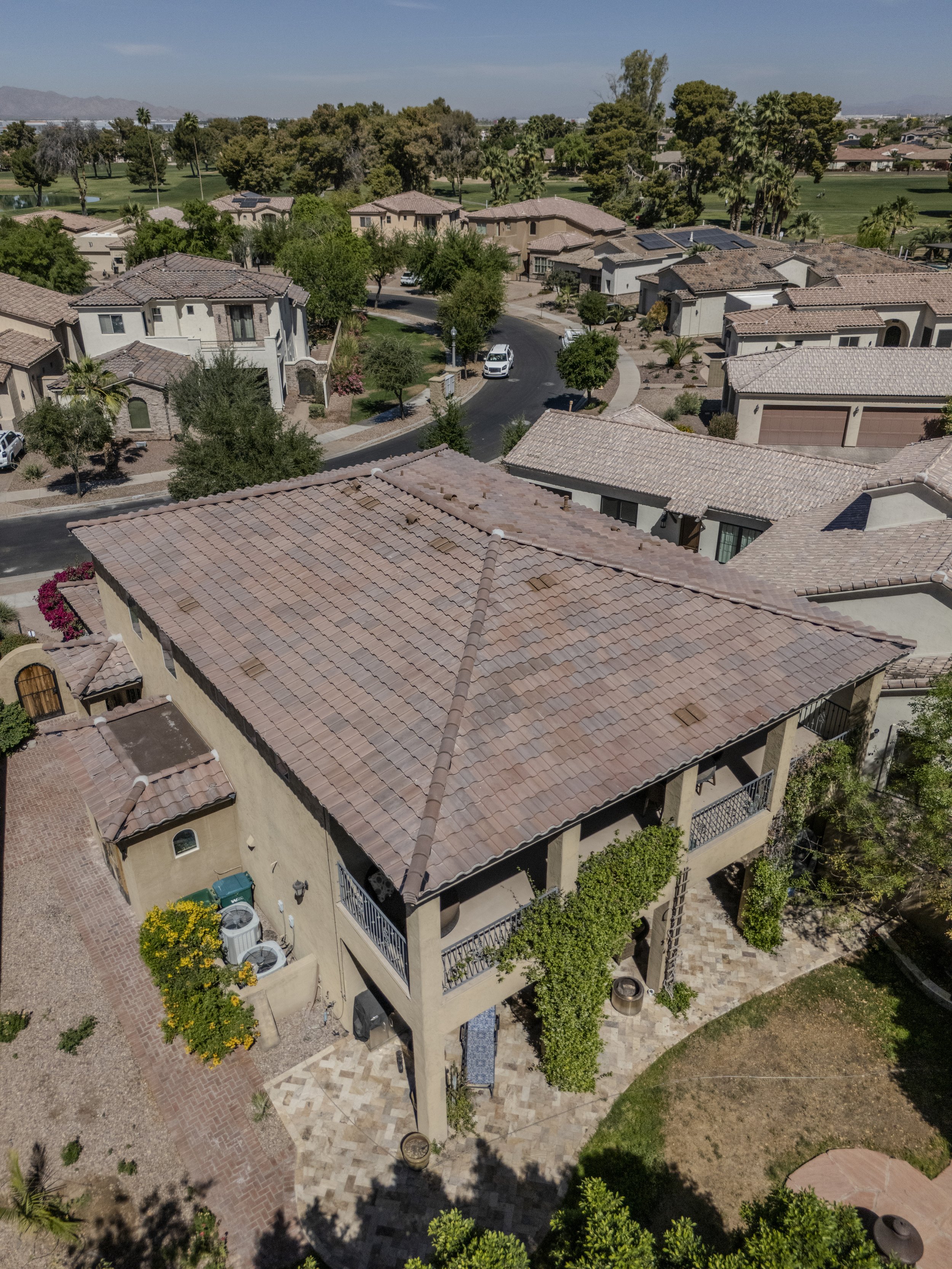 Aerial view of a suburban neighborhood with houses featuring beige and cream stucco exteriors and red-tiled roofs, surrounded by greenery, trees, and a park in the background.