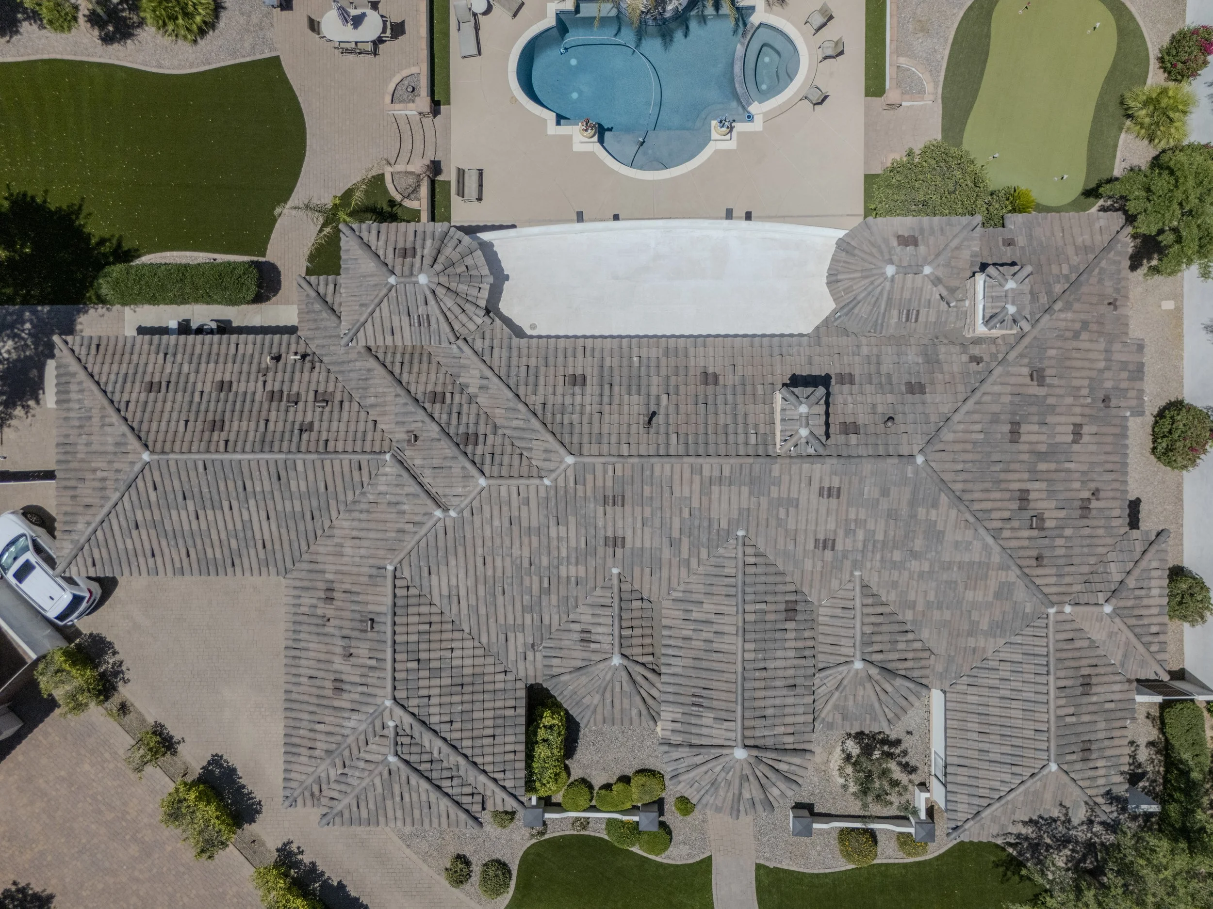 Aerial view of a backyard with a swimming pool, surrounded by a paved patio area, lawn, trees, and neighboring rooftops.