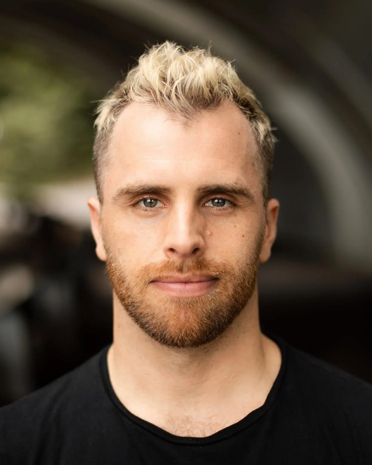 A young man with blonde, curly hair and a beard looks directly at the camera. He is wearing a black shirt and the background is blurred.