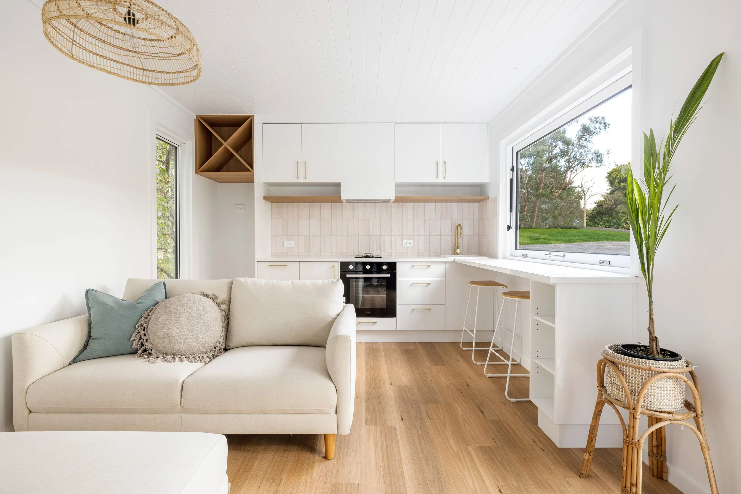 Interior of a modern, minimalistic kitchen with white cabinets, wooden accents, large window showing outdoor greenery, and a cozy white sofa with pillows in the foreground.