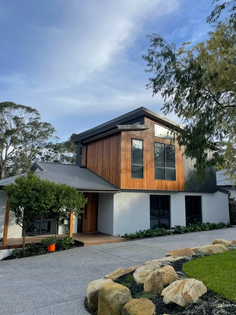 A modern two-story house with a mix of white and wooden exterior, large windows, surrounded by a landscaped yard with rocks, grass, and trees, under a partly cloudy sky.