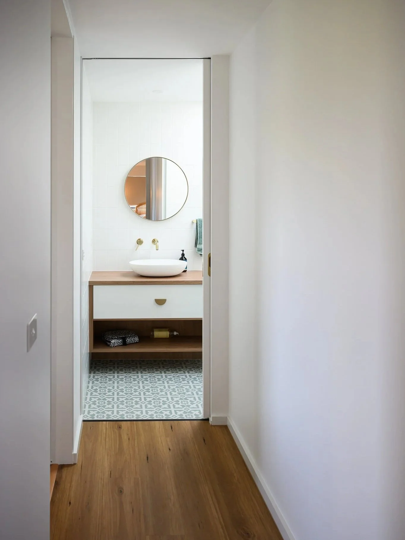 A view into a small bathroom with a white vessel sink on a wooden vanity, a round mirror above, and patterned floor tiles, seen through a doorway with the main room's wooden floor and white walls.