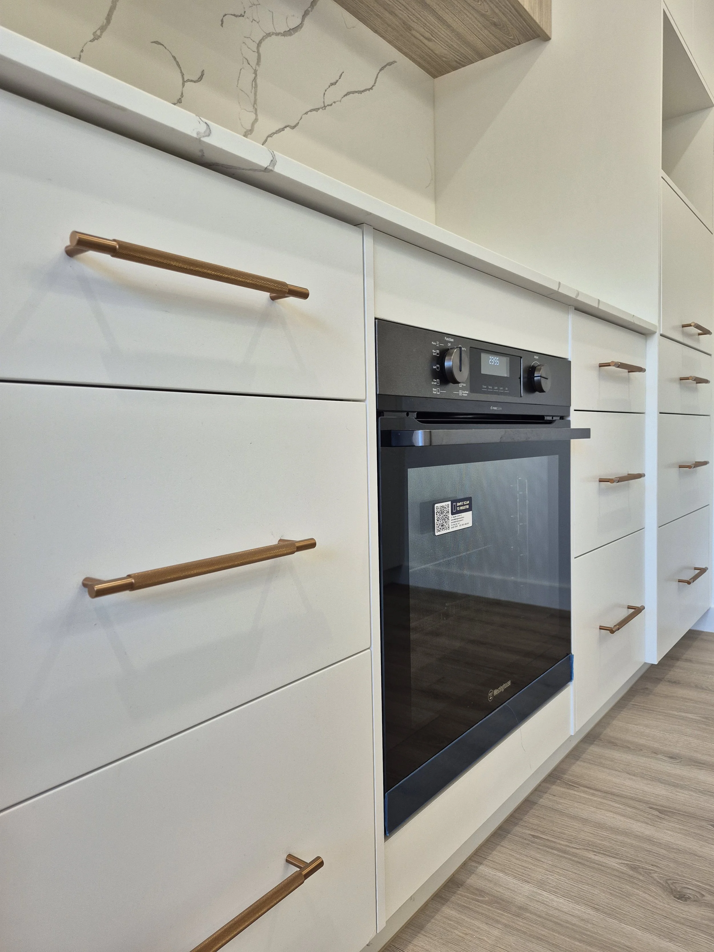 Modern kitchen cabinets with white finish and gold handles, a built-in black oven, and light wood flooring.