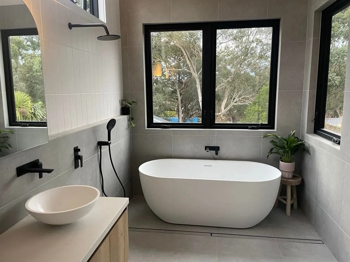 Modern bathroom with large bathtub, black fixtures, a bowl sink, and large windows showing trees outside.