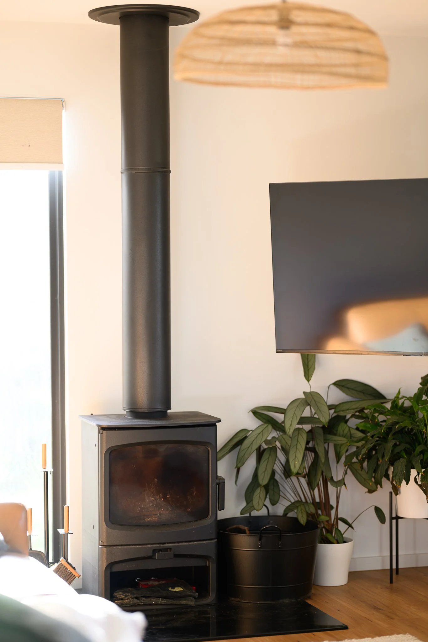 A cozy living room with a black wood stove, a chimney pipe extending to the ceiling, a small wood storage drawer, a potted plant with large green leaves, a black indoor plant container, a white pot, and a wooden floor. There is a lamp with a woven shade on a black floor lamp stand and a wall-mounted TV.