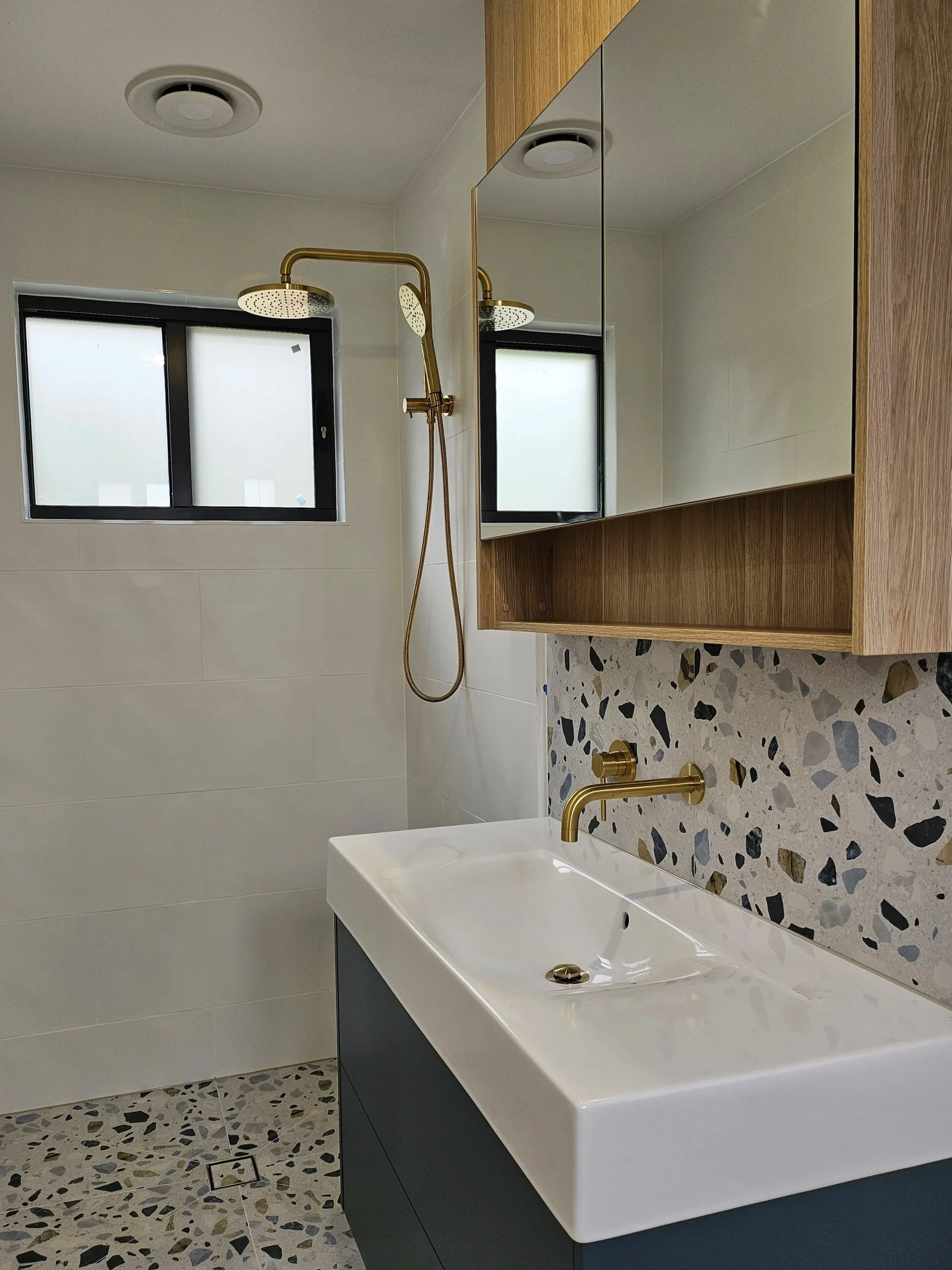 Modern bathroom with terrazzo floor, white walls, a black window, a white porcelain sink with a gold faucet, and a wooden-framed mirrored cabinet.