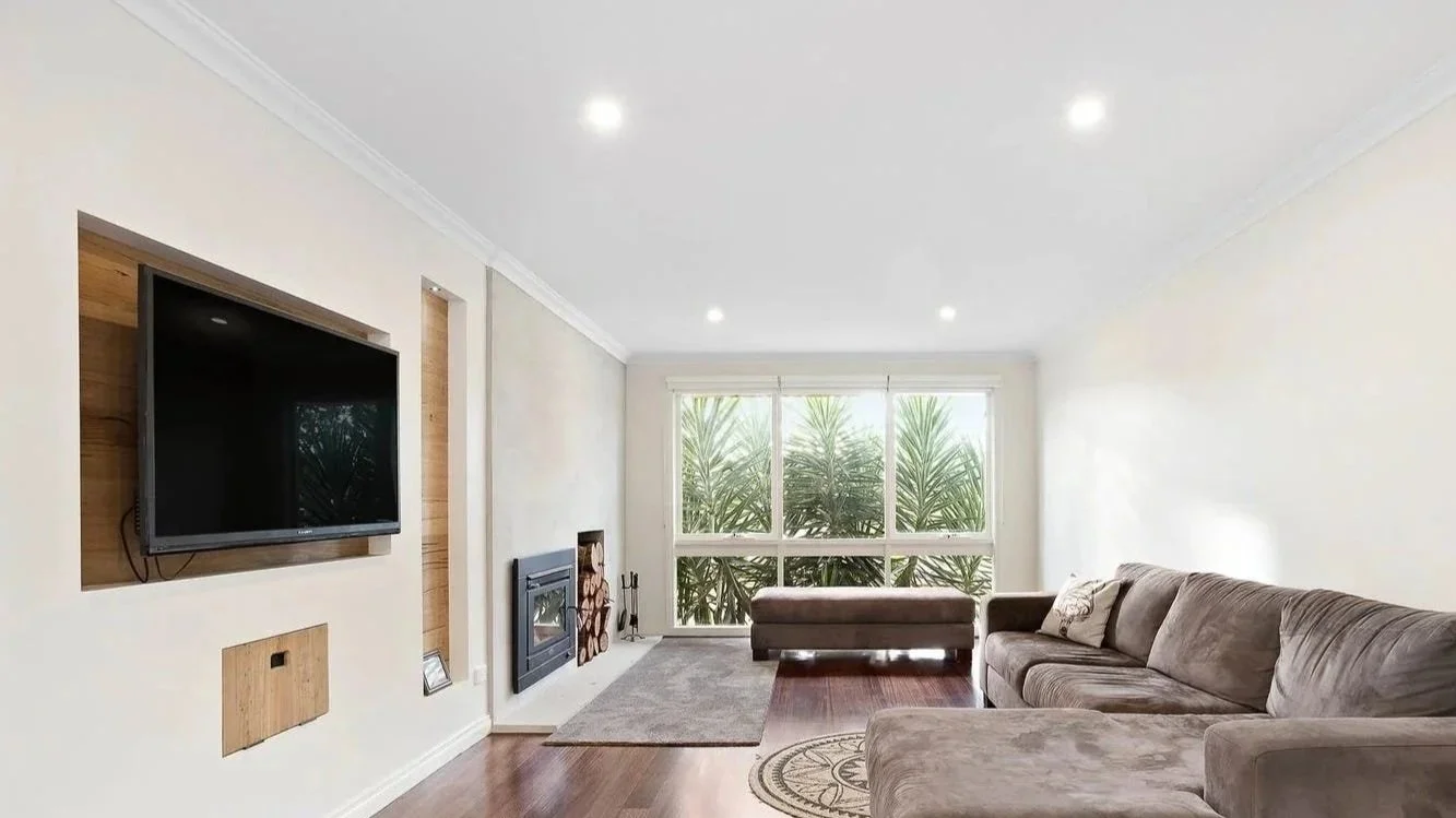 Living room with a wall-mounted TV, fireplace, large window with greenery outside, beige sofa, and a small ottoman.