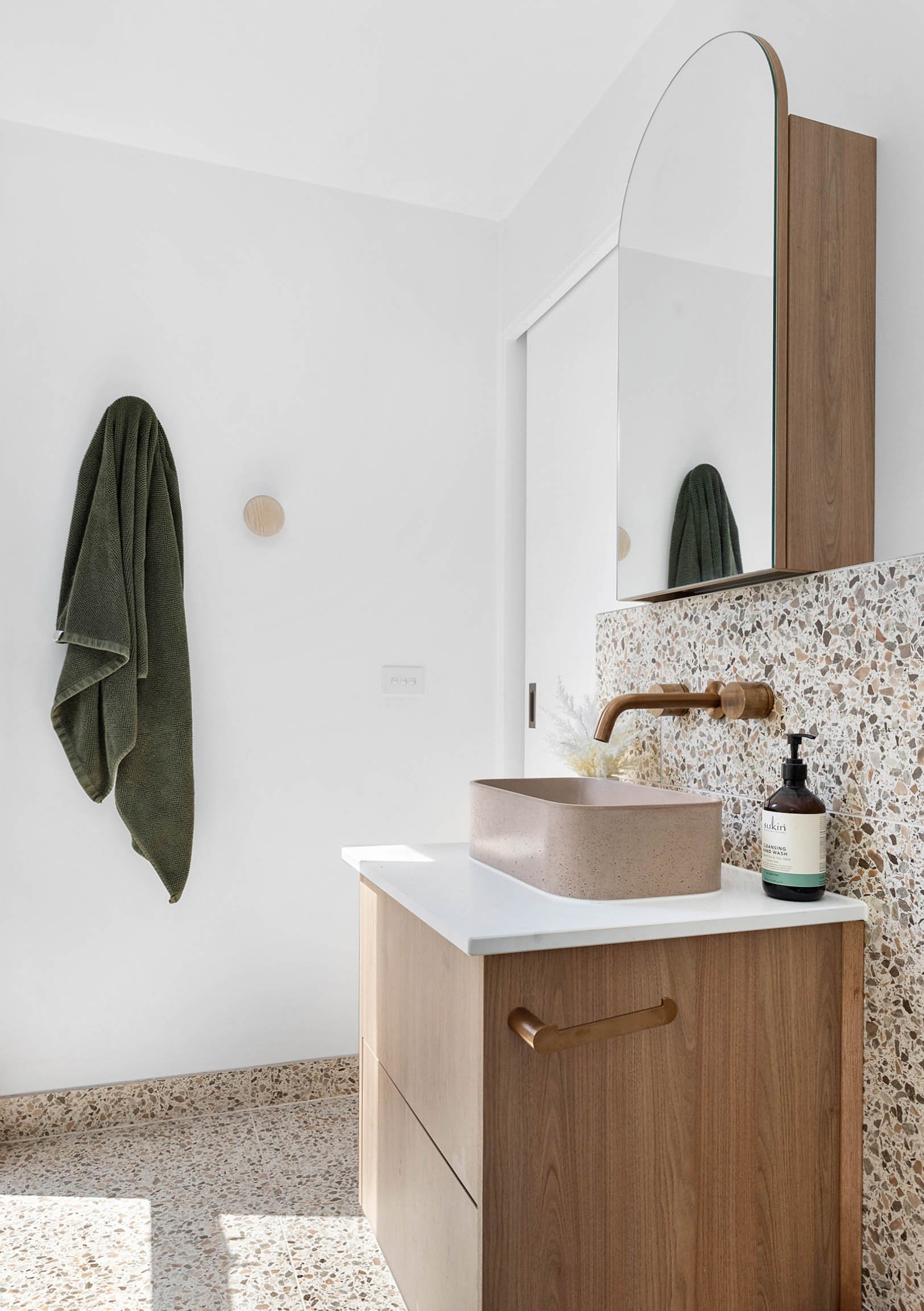 Modern bathroom with a terrazzo countertop, a round concrete sink, a wooden vanity with drawers, a mirror, a greenery soap dispenser, and a green towel hanging on the wall.