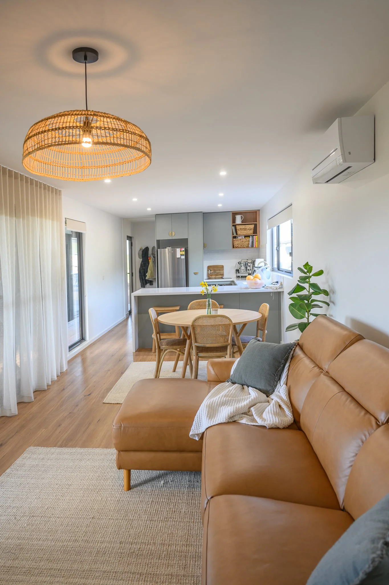 Open-plan living area with a tan leather sofa, a wooden dining table with chairs, a kitchen island, white cabinets, and an air conditioning unit on the wall. There is a window with white blinds and a sliding glass door with sheer curtains.