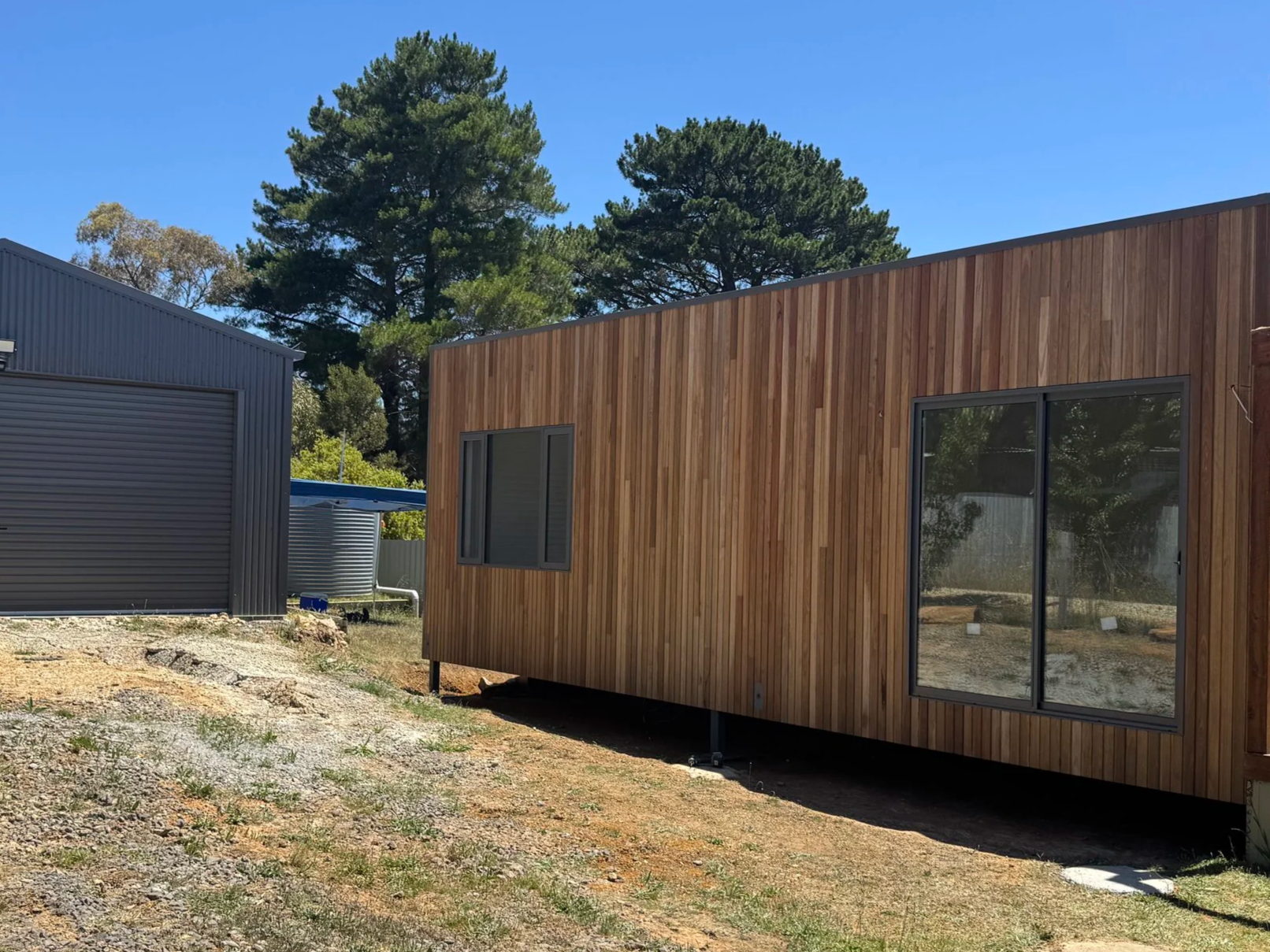 A modern wooden house with large windows next to a gray metal shed, situated outdoors with trees and a clear blue sky in the background.