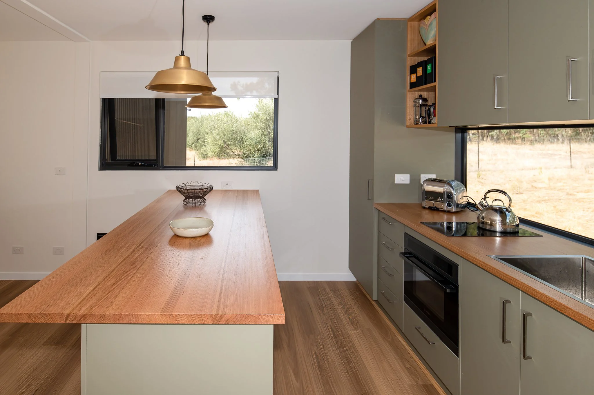Kitchen with a wooden island, gray cabinets, stainless steel appliances, open shelves, and two pendant lights hanging from the ceiling. Windows offer a view of greenery outside.