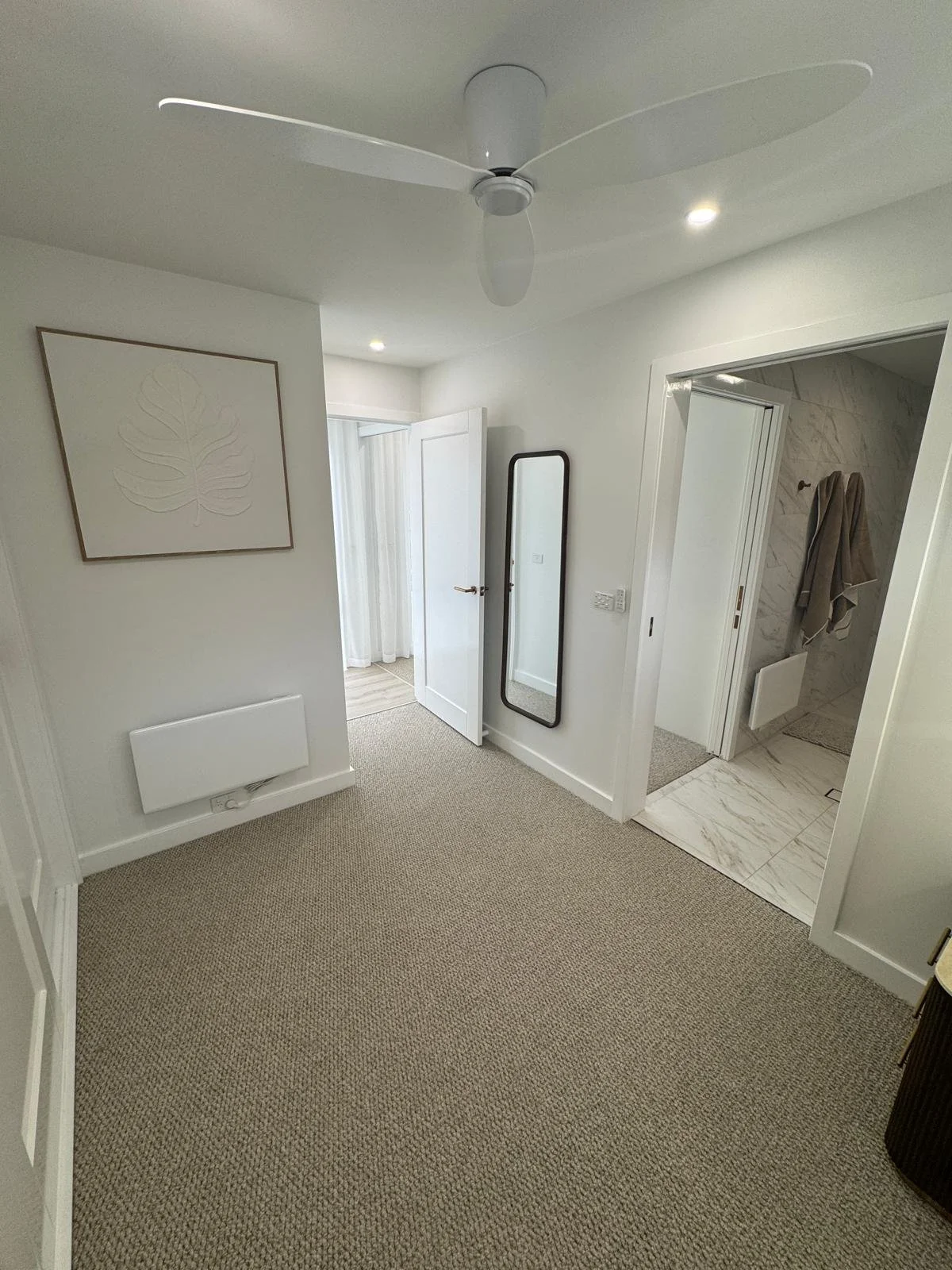 A clean, minimalist bedroom with a ceiling fan, framed wall art of a leaf, a full-length mirror, and an adjoining bathroom with marble tiles and towels.