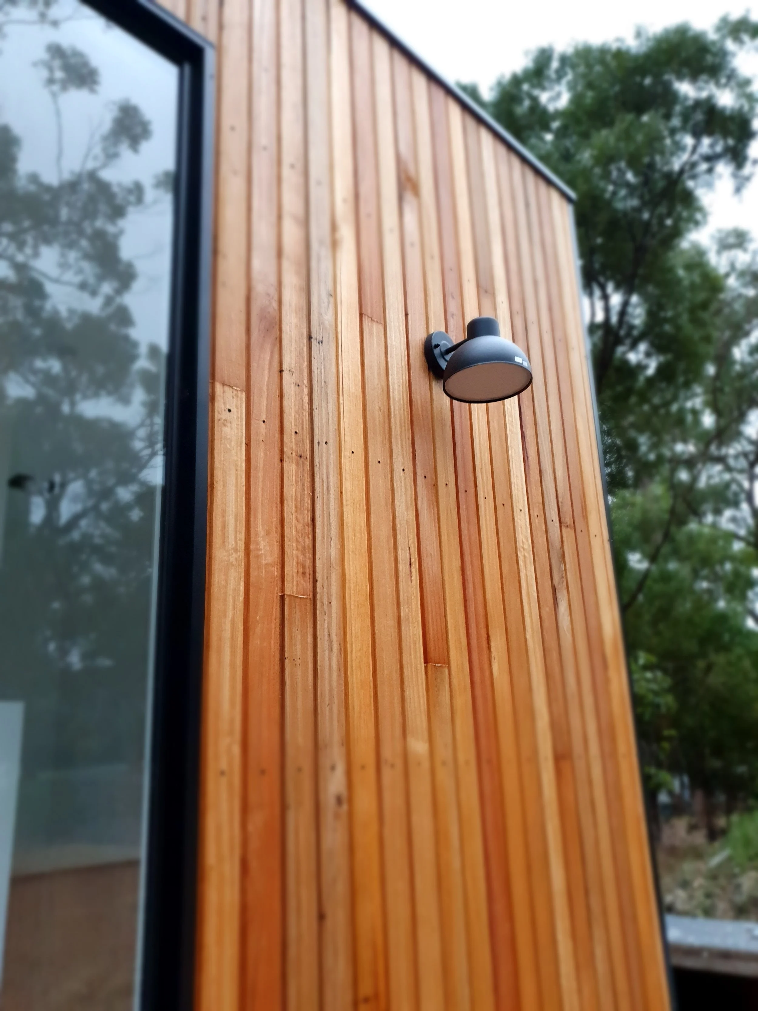 Exterior view of a building with wooden vertical siding and a black outdoor wall-mounted lamp, with trees and sky in the background.