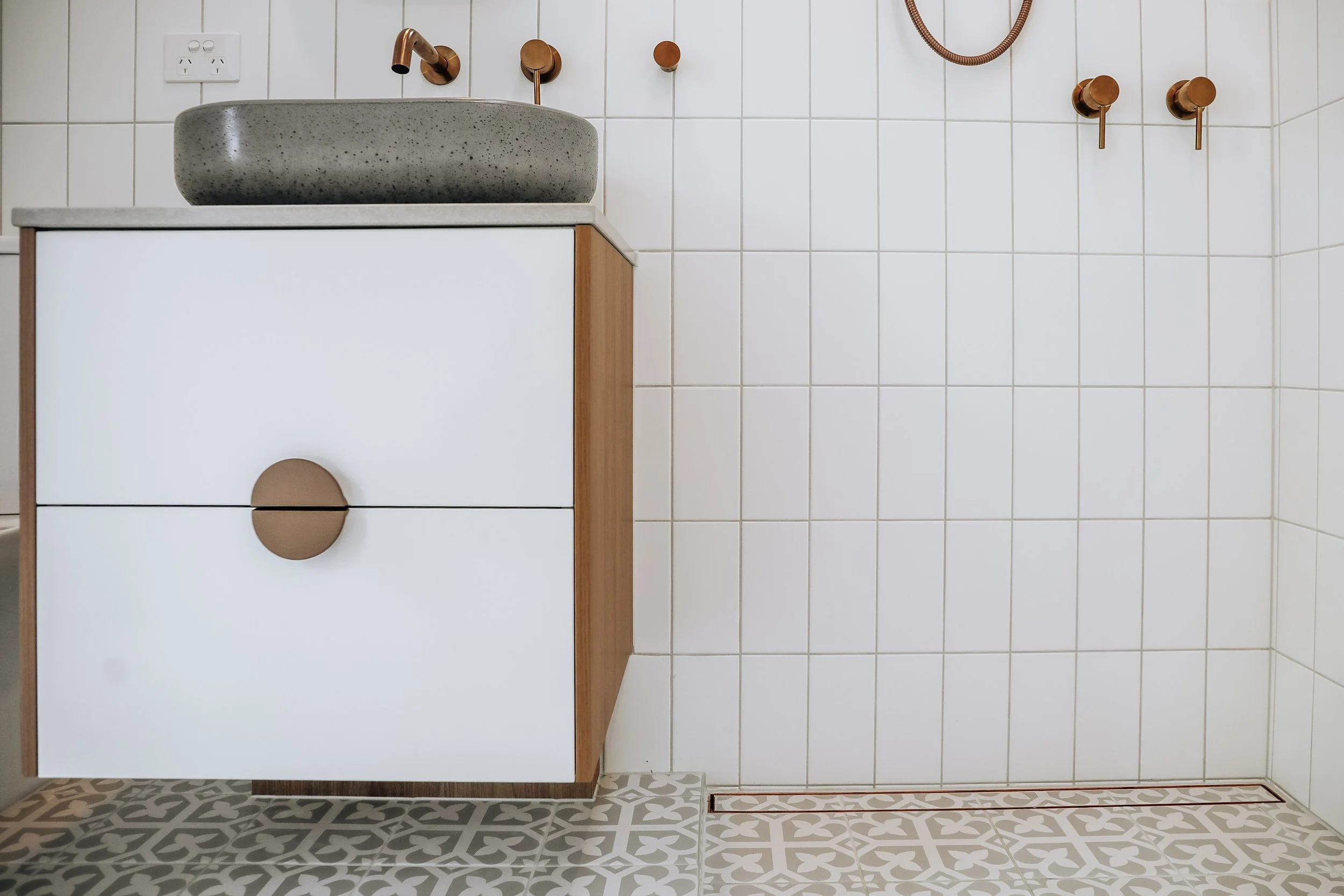 A bathroom vanity with a stone sink, copper fixtures, and a wooden framed cabinet, white tiled wall, patterned floor tiles, and an electrical outlet.