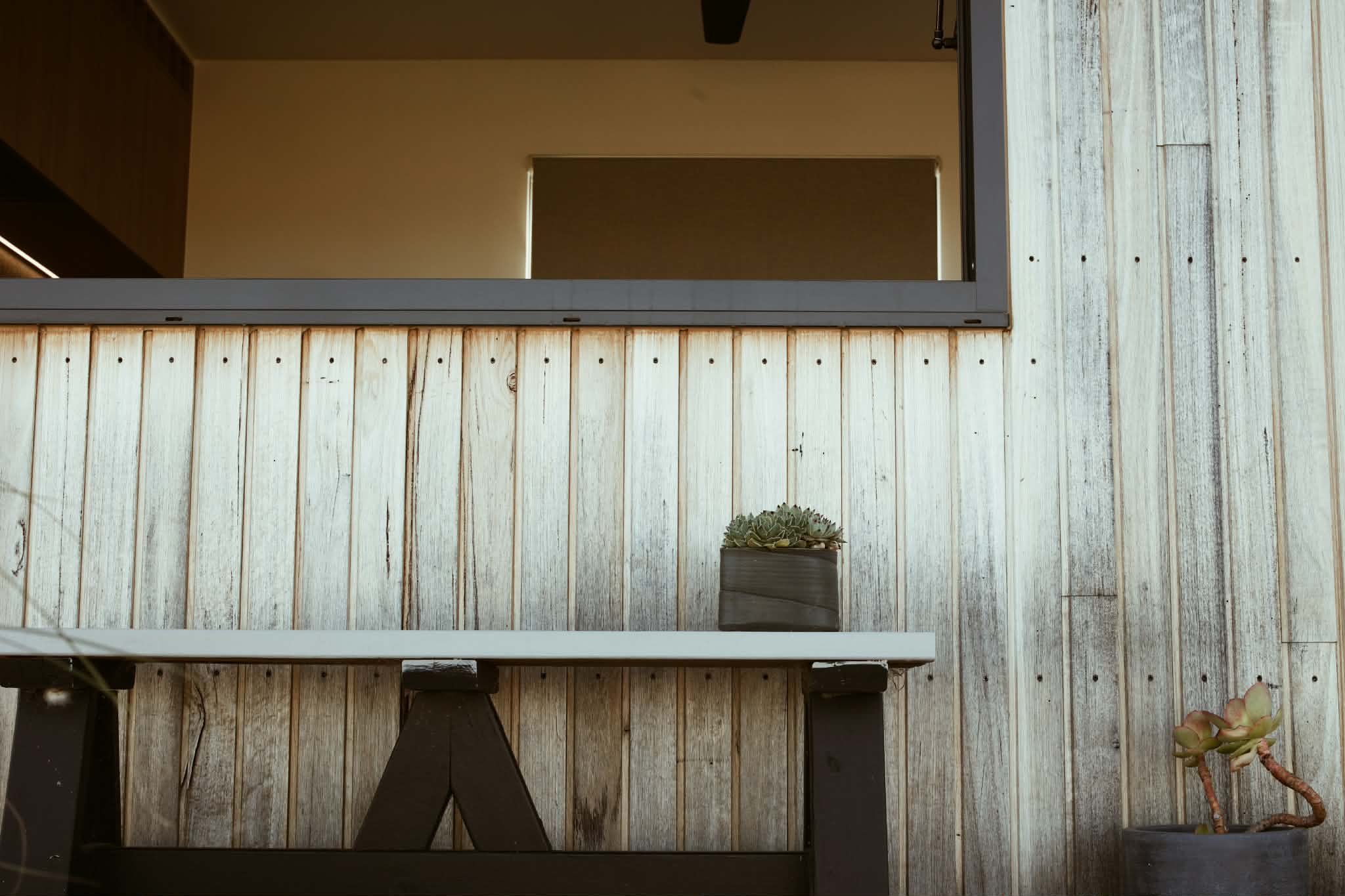 A view of a wooden deck with a succulent plant in a gray pot on the right side and a dark table leg on the bottom left. The background shows a beige wall with a small door or window opening and a black ceiling fan.