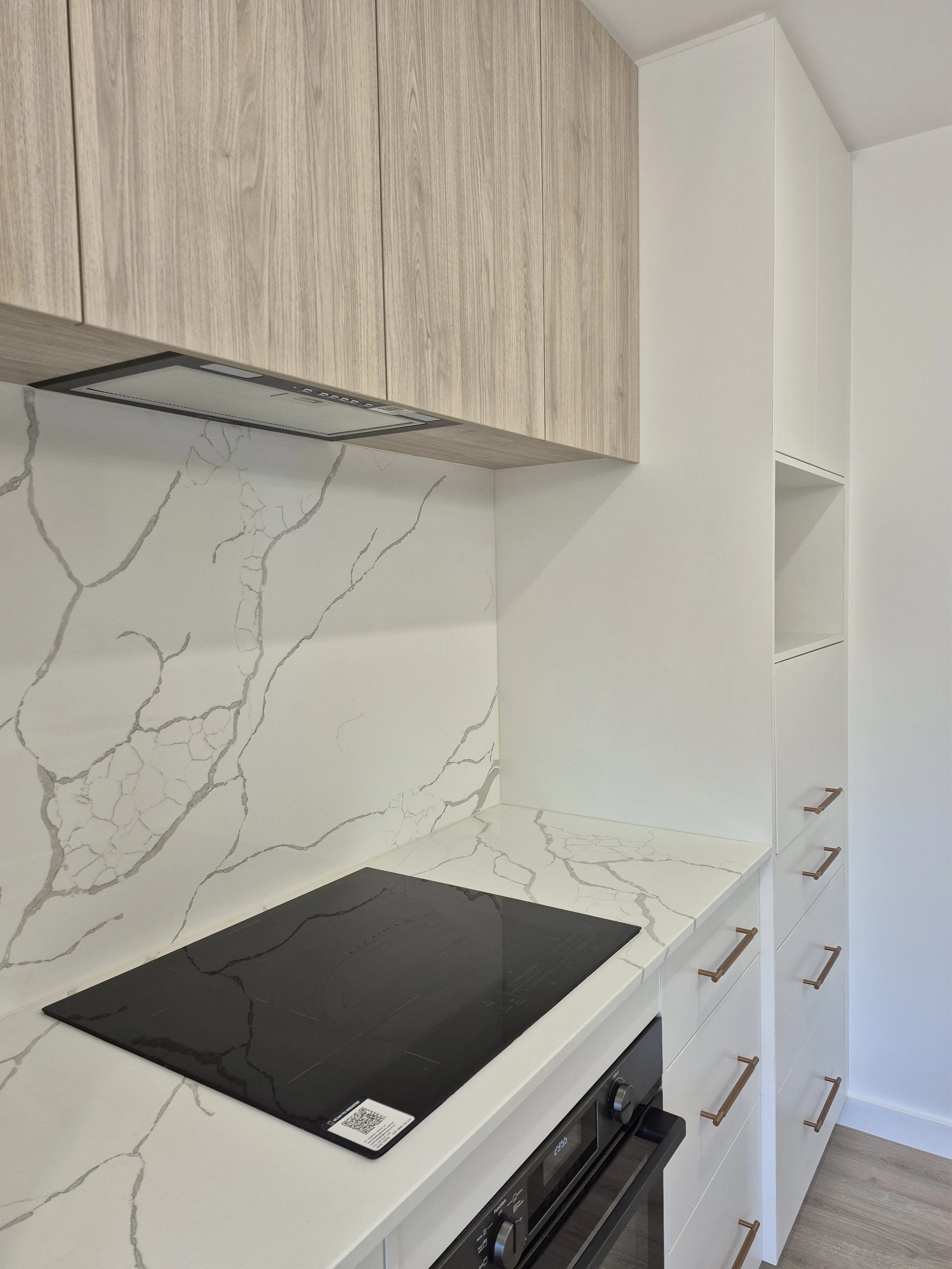 A modern kitchen countertop with a black electric cooktop, white marble backsplash with gray veins, and white cabinets with copper handles.