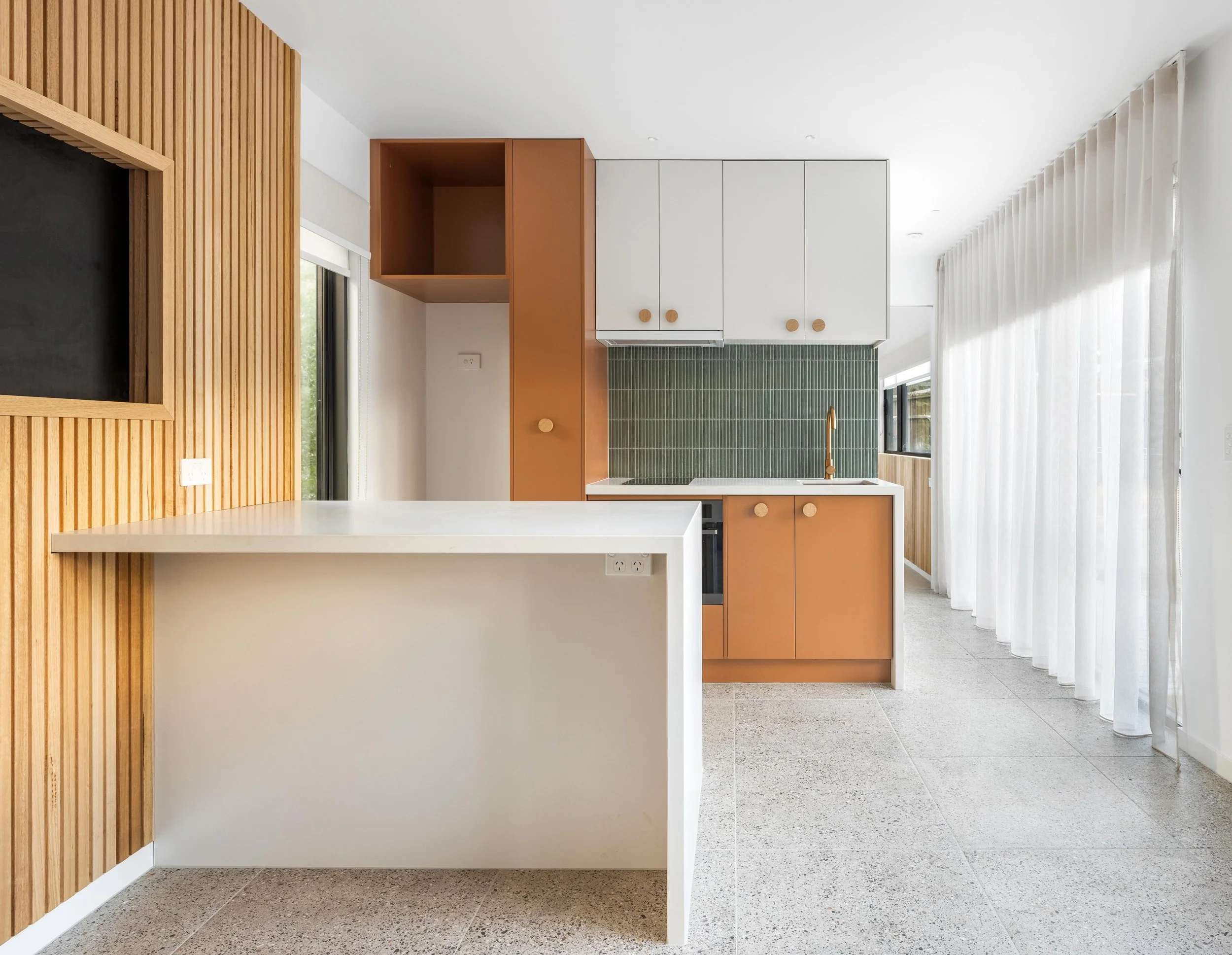 Modern kitchen with white cabinets, green subway tile backsplash, and a gold faucet, adjacent to a living space with wooden wall paneling, large windows with white curtains, and ceiling lights.