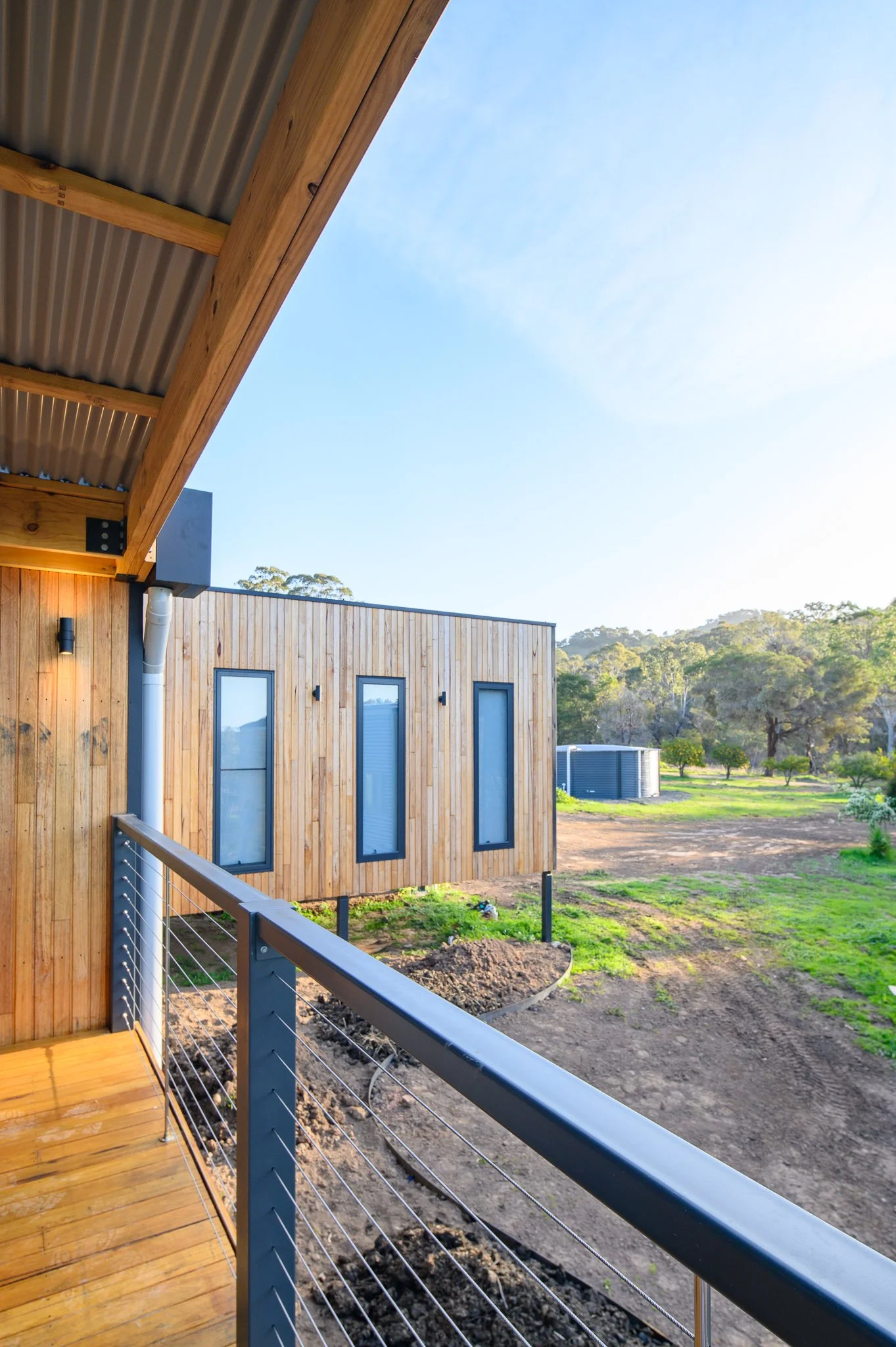 View from a wooden balcony with a metal railing overlooking a backyard with a new house. The house has wooden siding, tall narrow windows, and a sloped roof with a corrugated metal soffit. The yard has patches of grass, a garden bed, and a water tank, with trees and hills in the background under a blue sky.