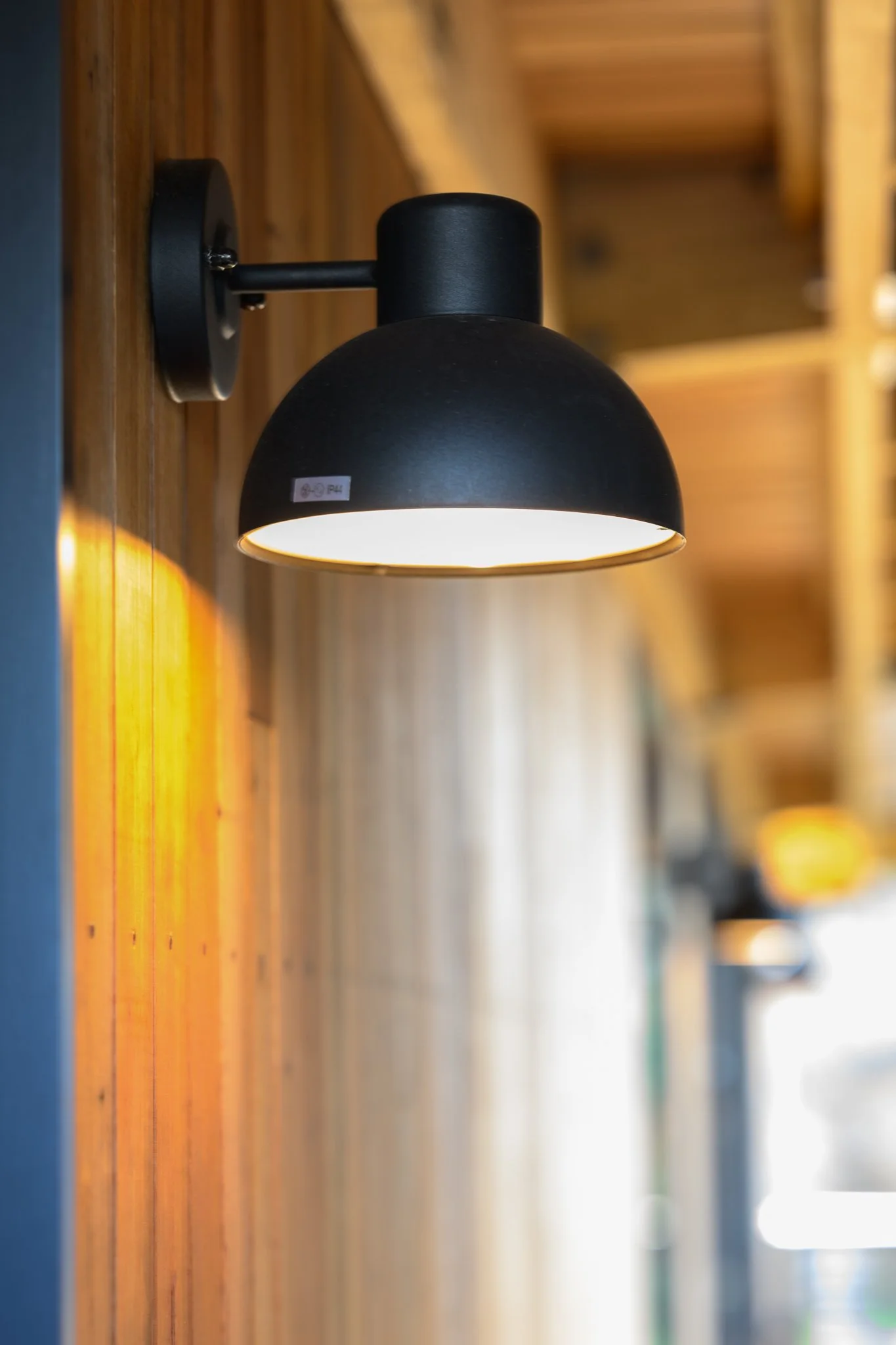 A black wall-mounted sconce light fixture attached to a wooden wall, with additional similar fixtures blurry in the background.