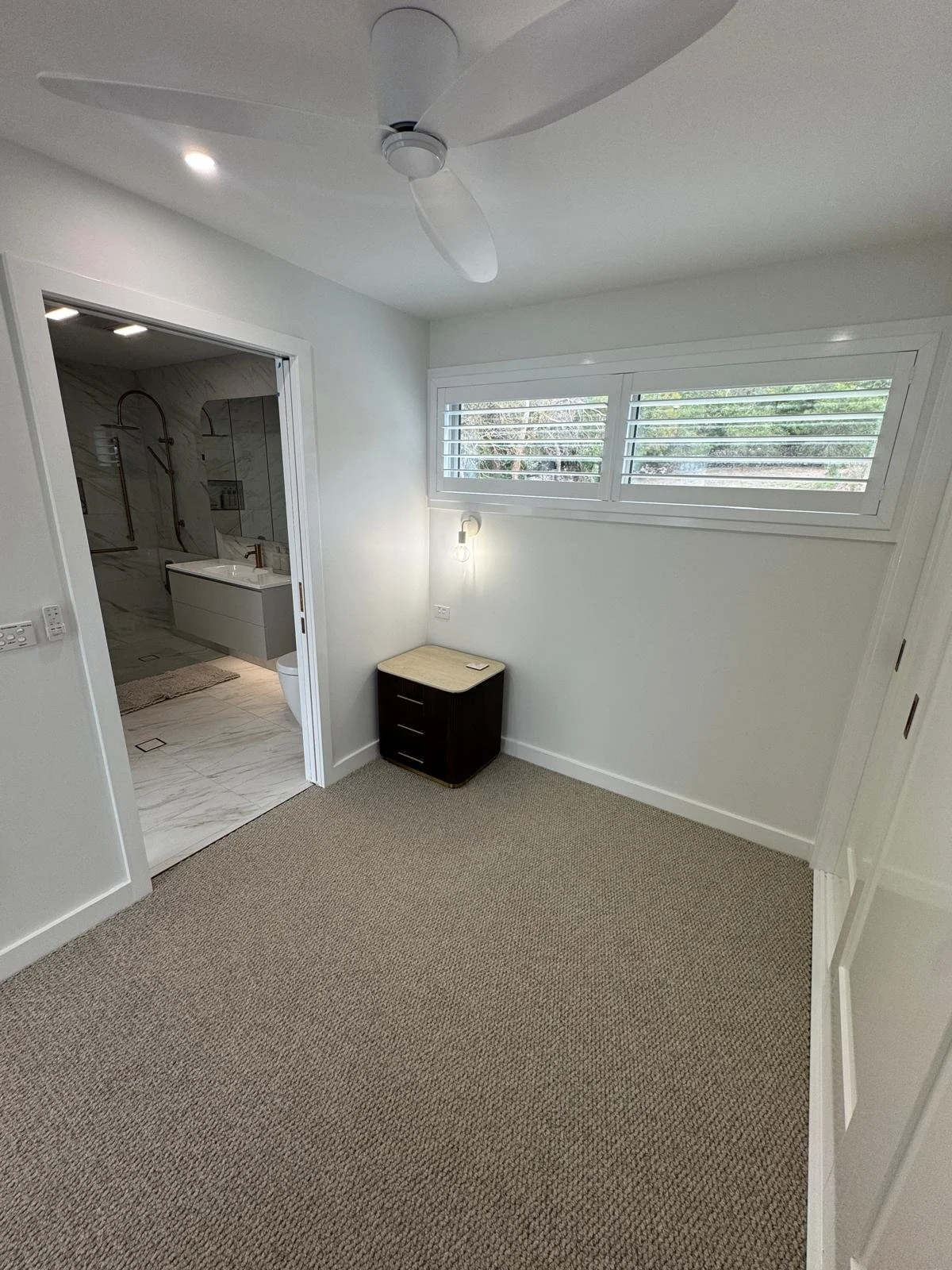 Empty room with a window, ceiling fan, small side table, and an open door leading to a bathroom with marble tiles and fixtures.
