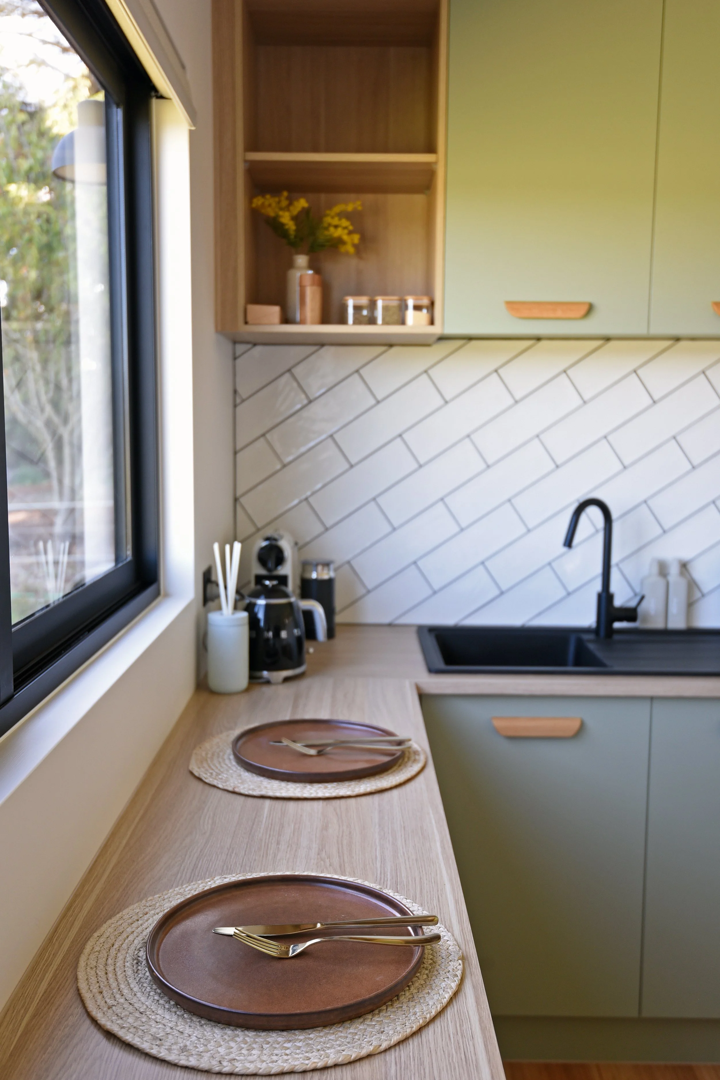 A kitchen with a wooden countertop, green cabinets, white tiled backsplash, and a large window showing outdoor greenery. The counter has a brown plate, silverware, and place mats, with a small appliance and reed diffuser nearby.