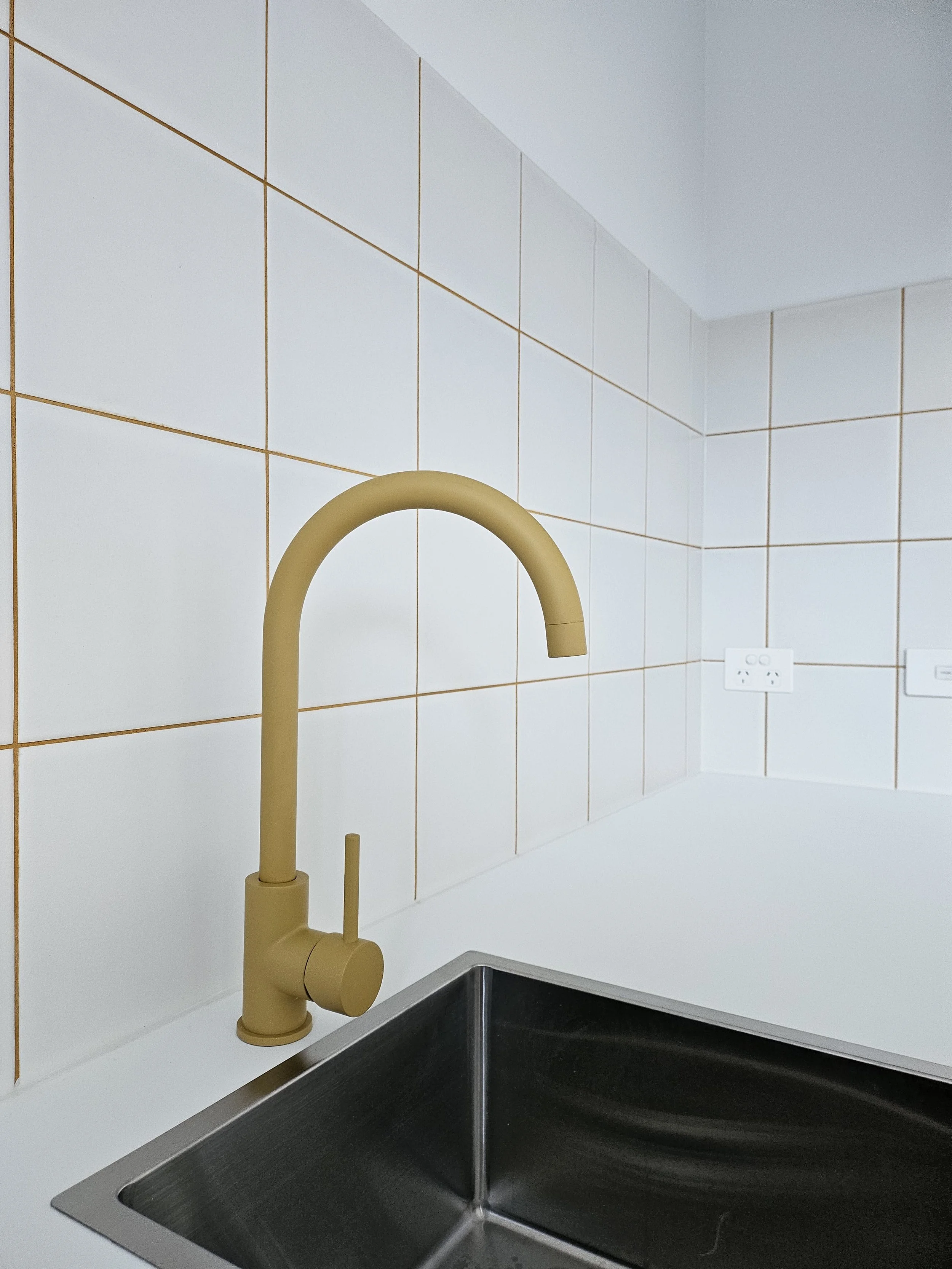 A kitchen sink with a black basin, beige faucet, white tiled wall, and a white electrical outlet.