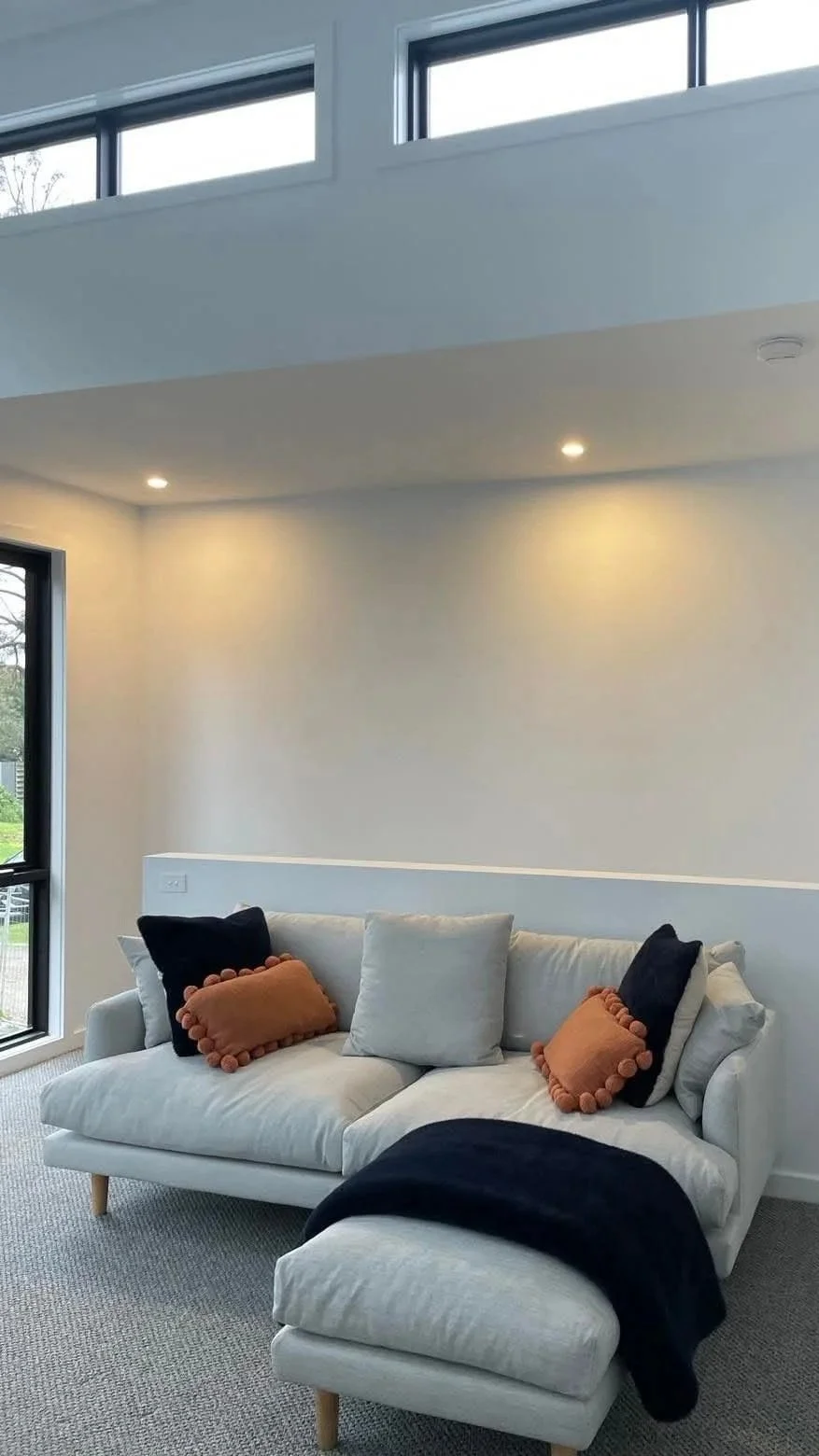 Modern living room with white sofa, black and orange throw pillows, a black throw blanket, large windows, and recessed ceiling lights.