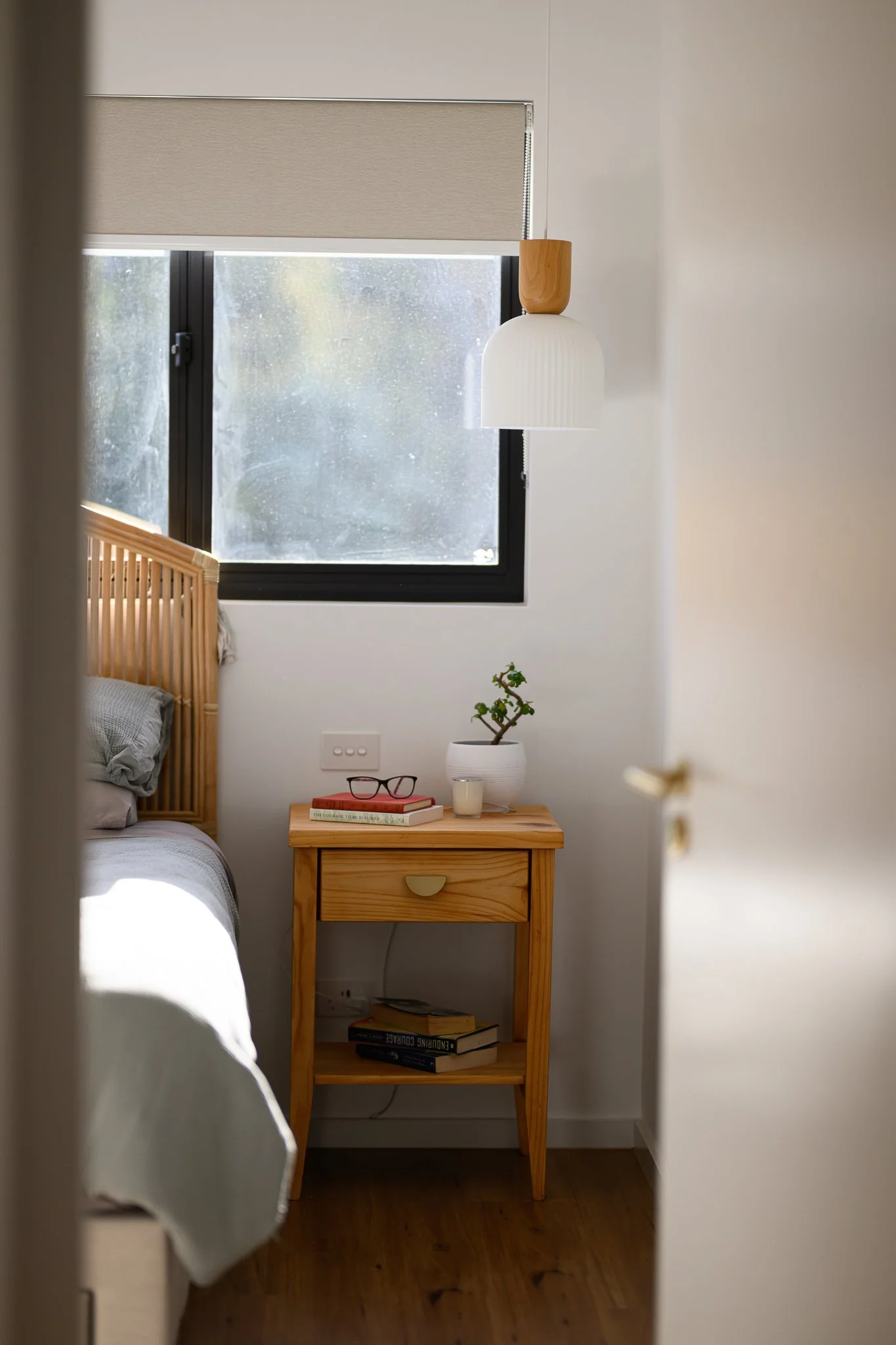 A glimpse of a cozy bedroom with a wooden nightstand, a small potted plant, books, a candle, and a pair of glasses. The bed has a wooden headboard and beige bedding, situated next to a window with a roller shade. The room has wooden flooring and a modern, minimalist design.