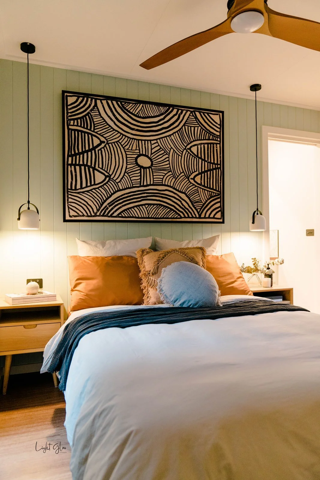 A cozy bedroom with a bed featuring cream, peach, and light blue pillows, a beige comforter, and a black and gray striped throw blanket. Two matching wall-mounted pendant lights hang on either side of the bed, and a wooden nightstand with books and decor is on the left. A large, black and white abstract art piece hangs above the bed, with a ceiling fan above and a window on the right allowing natural light.