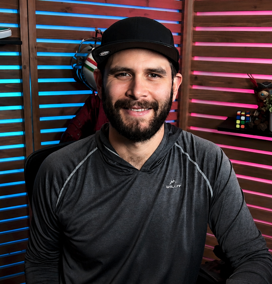 A man with a beard wearing a black cap and dark athletic shirt, smiling in front of a colorful, wooden slat background with blue and pink lighting.