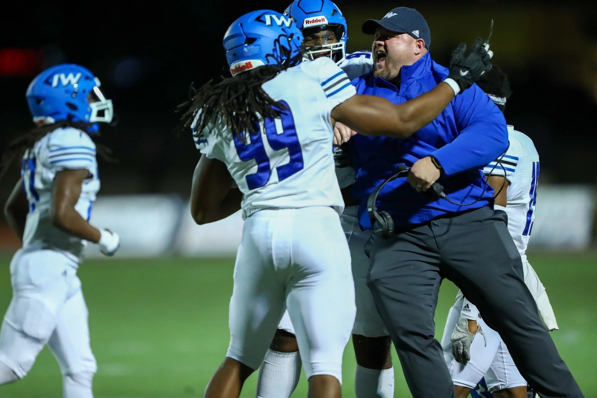 American football coach celebrates with a player on the field during a game, with players wearing blue helmets and white uniforms.