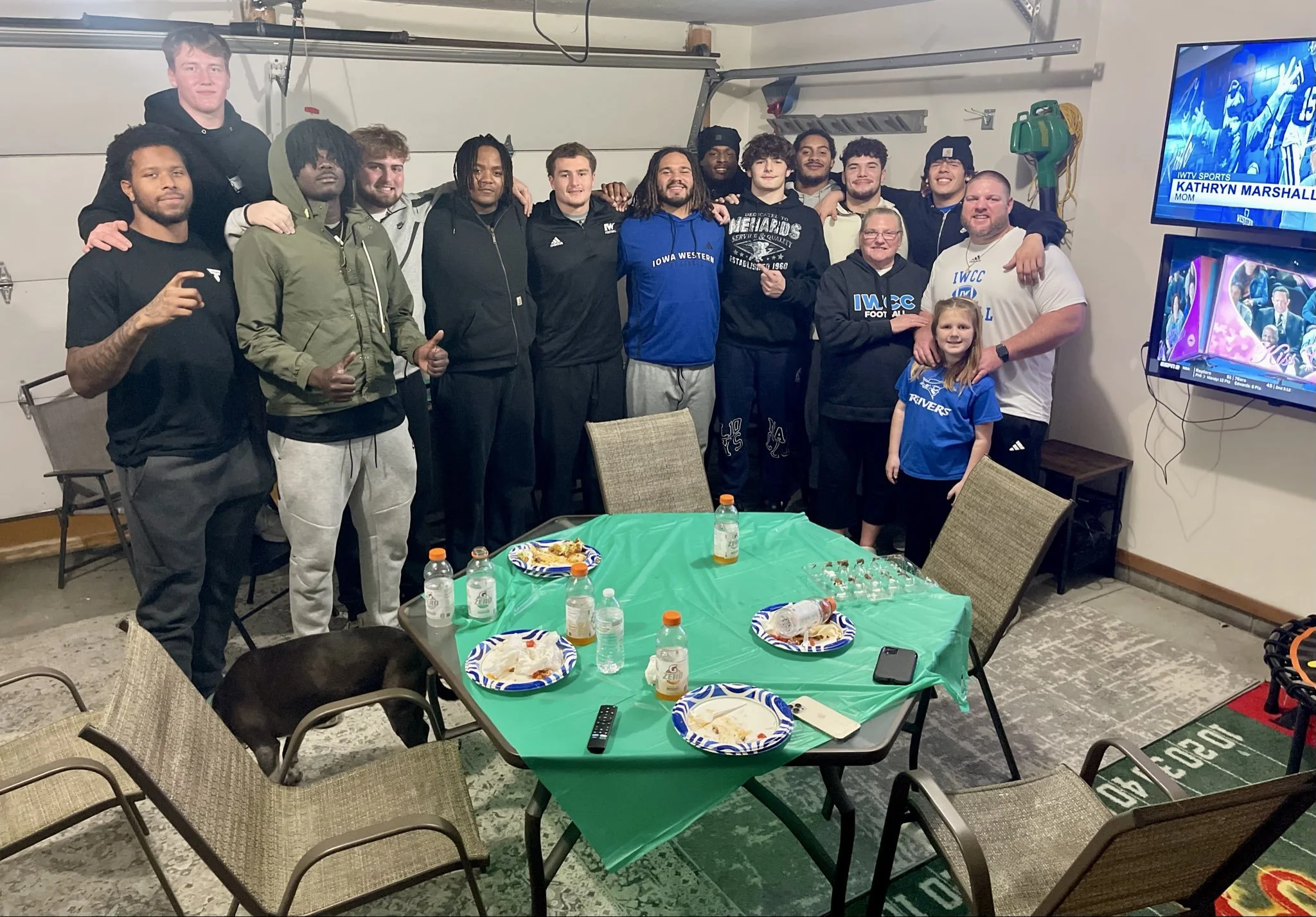 Group of people, including young men and an older woman, posing together in a garage decorated with sports memorabilia, celebrating an event with food and drinks on a table. There is a television displaying sports news.