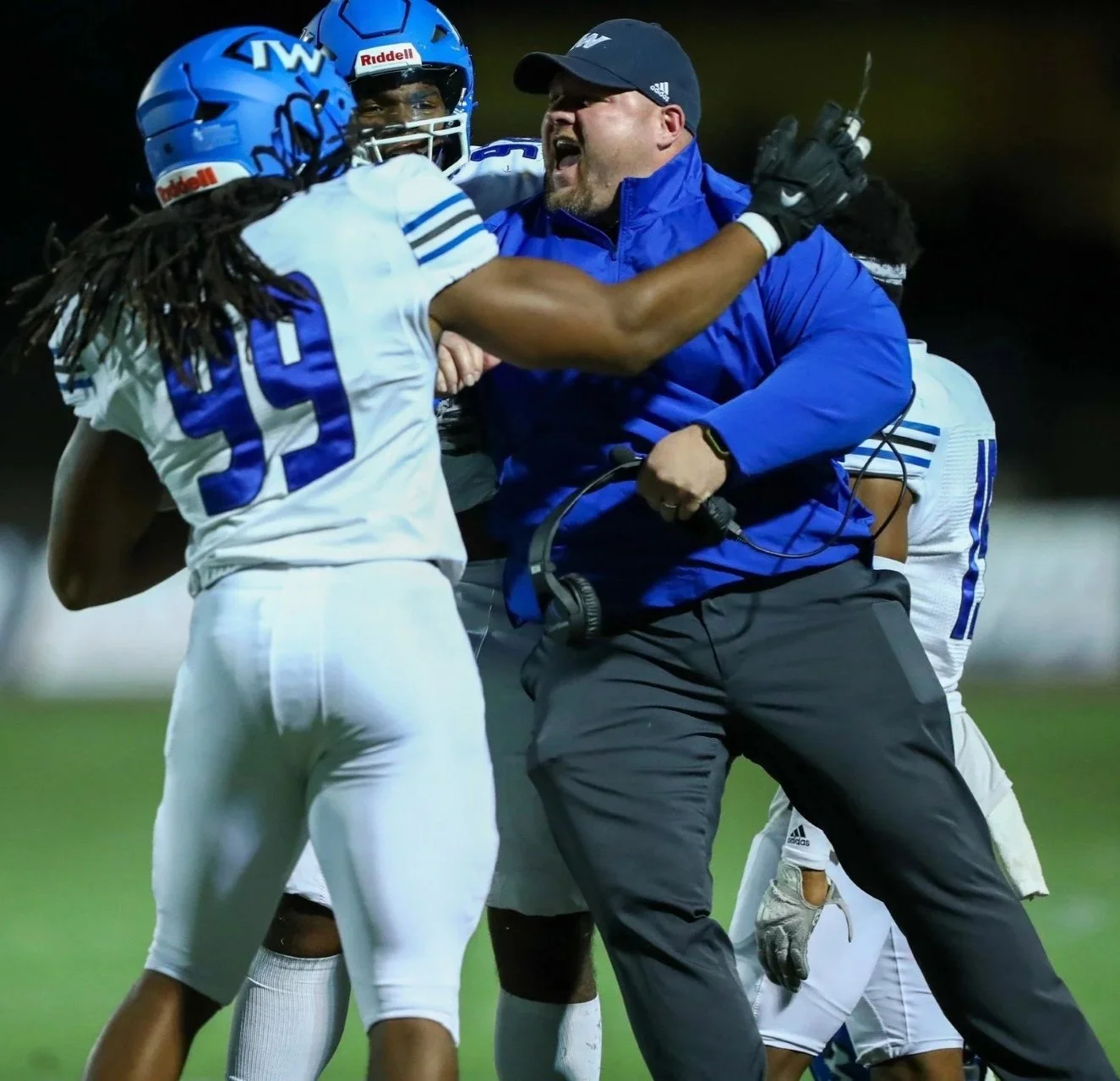 Football players and coach engage in a heated altercation on the field, with players wearing blue helmets and jerseys, and the coach shouting with a tense expression.
