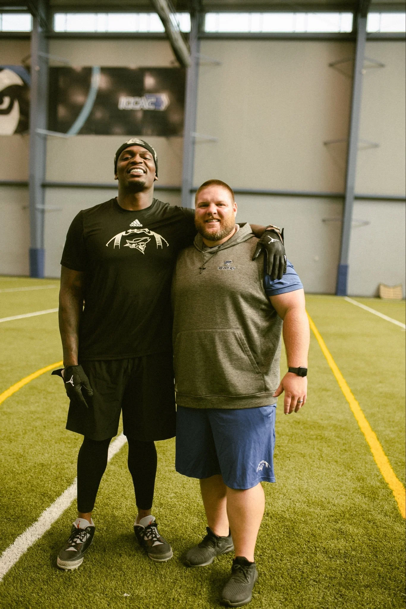 Two men standing together on an indoor sports field, smiling at the camera. The man on the left is wearing a black athletic shirt, black shorts, and sneakers, with gloves and a beanie. The man on the right is wearing a gray vest over a blue shirt, blue shorts, and sneakers, with a cross necklace and a watch.