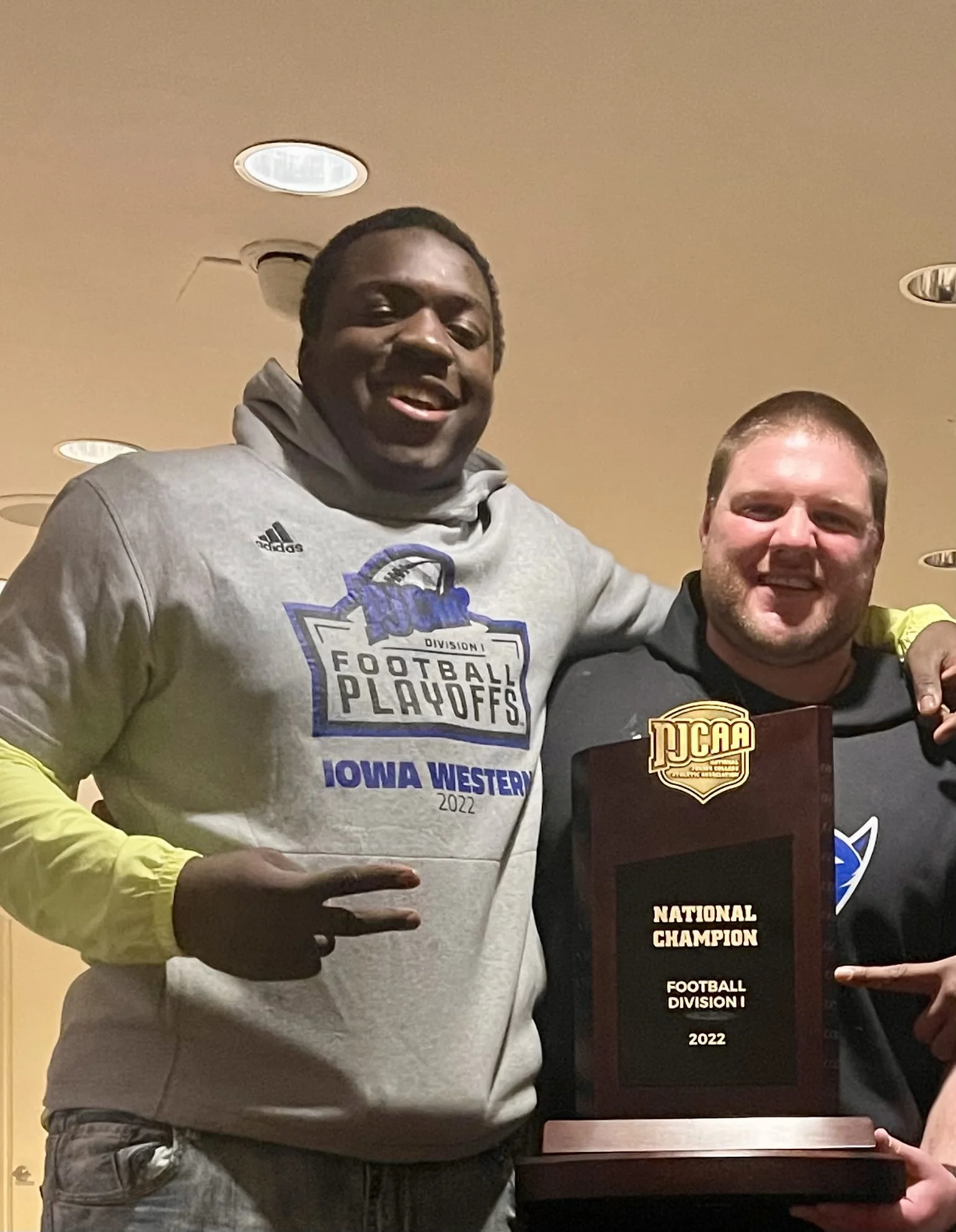 Two men smiling and holding a trophy for the football division 1 national champion at an indoor event, with ceiling lights above.