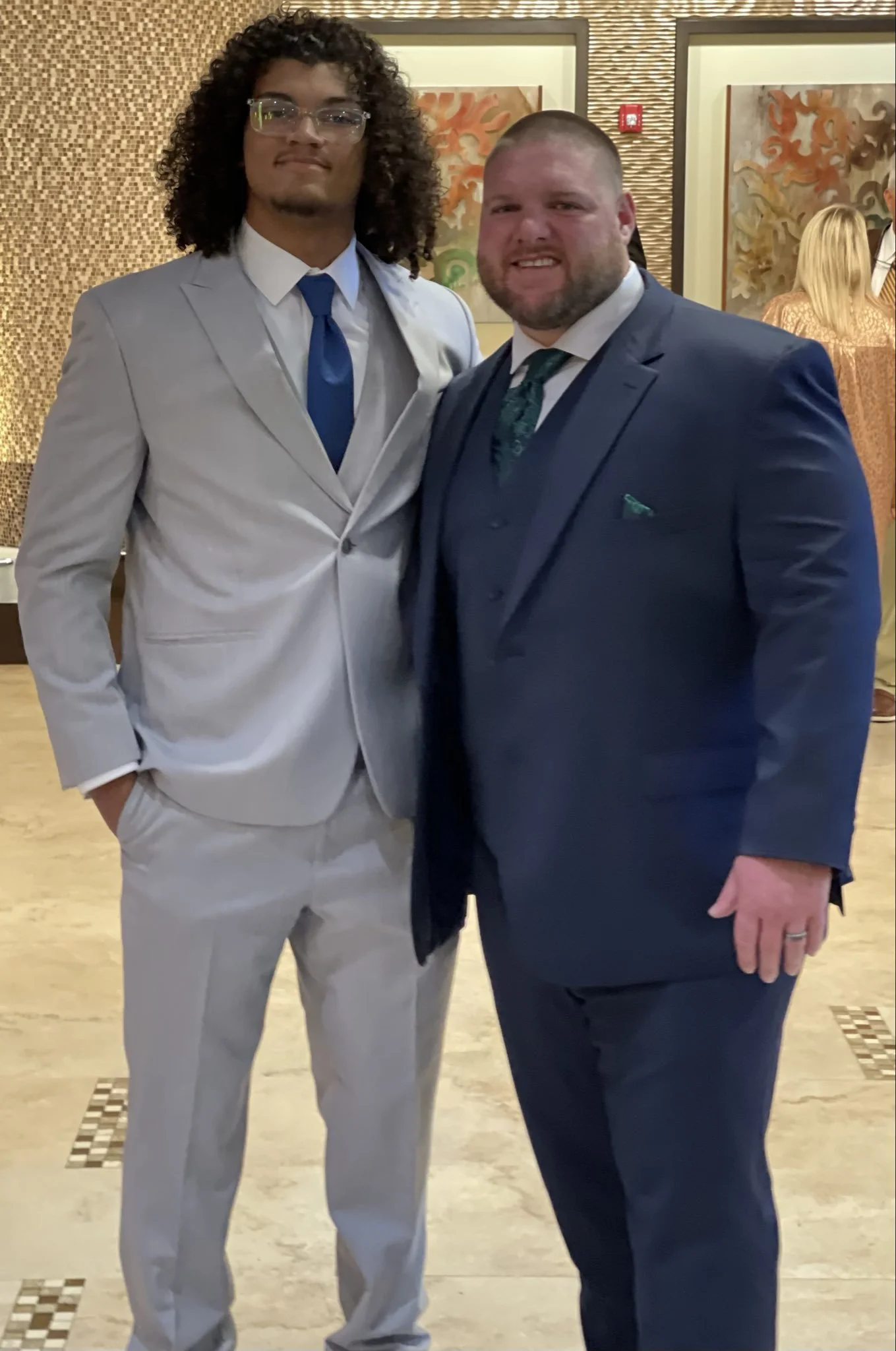 Two men dressed in suits posing together indoors, with artwork and other people in the background.